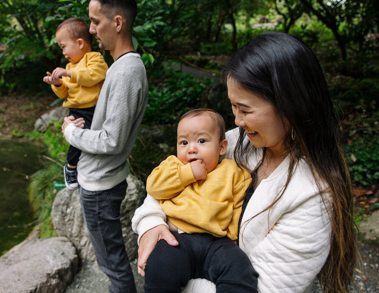 photo of Asian mom carrying baby son in yellow sweatshirt while dad holds toddler son in the background