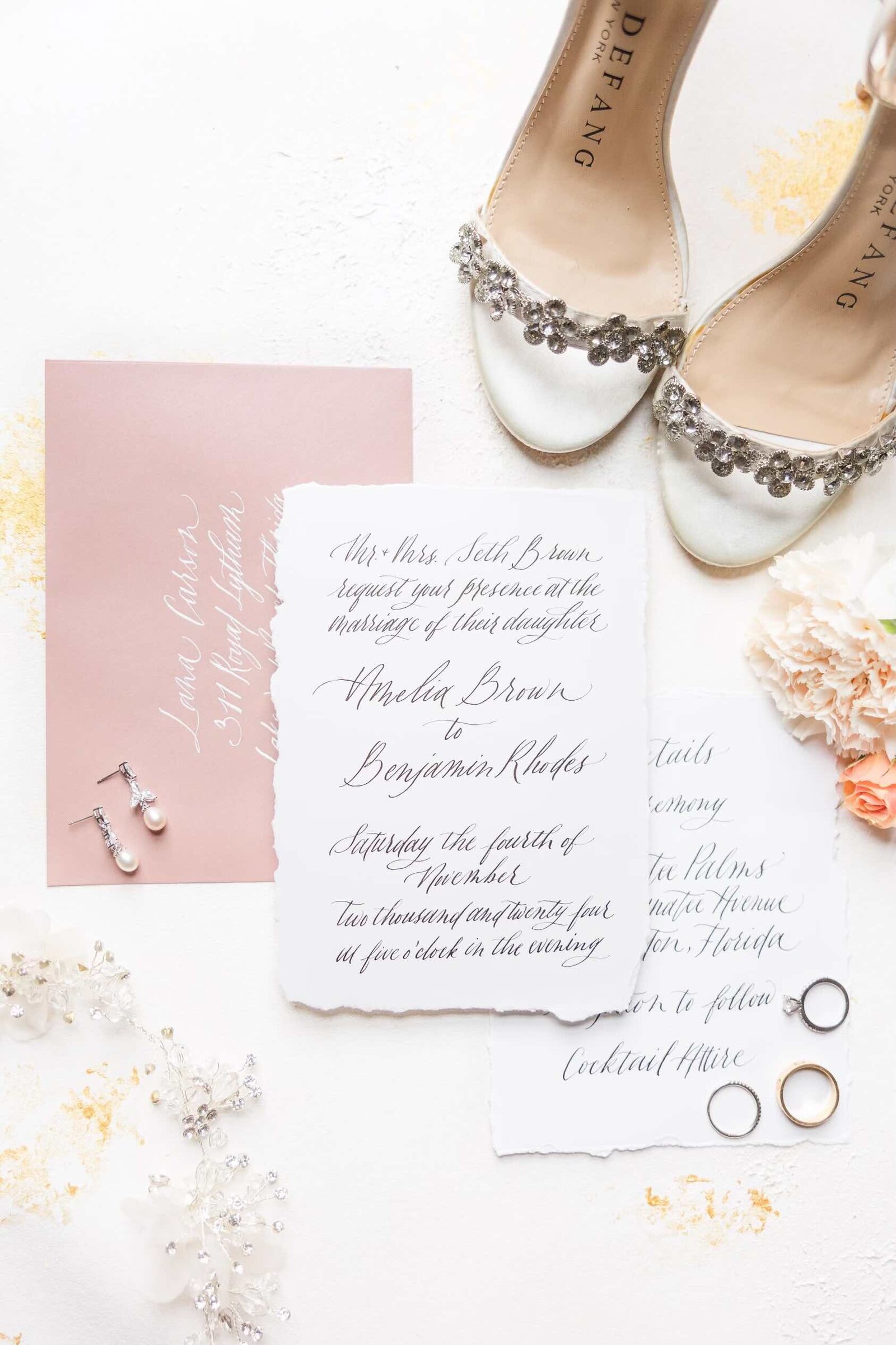 handwritten invitation suite, rings, wedding shoes, calligraphy envelope, and florals scattered around the invites
