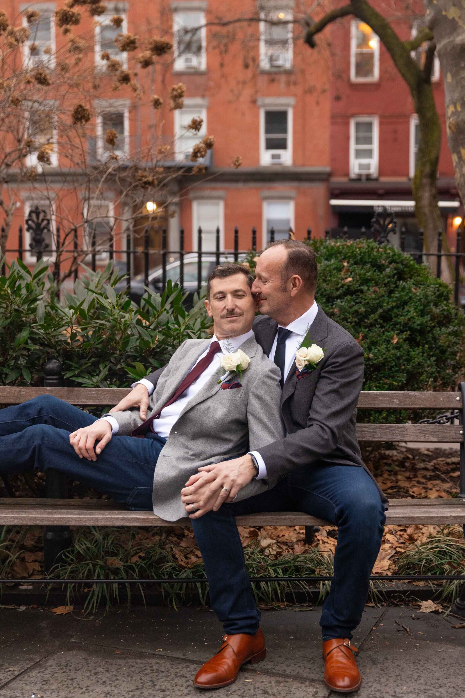 Two grooms sitting on a park bench leaning on each other.