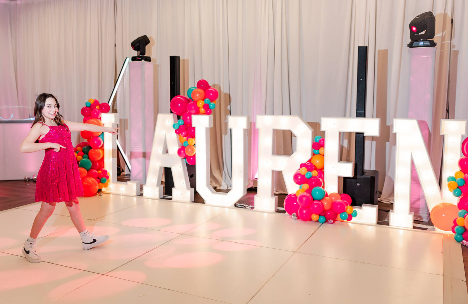 A teen girl in a bright pink dress points to her name in large light up letters on the dance floor during a Bellevue Bar and Bat Mitzvah Photography session