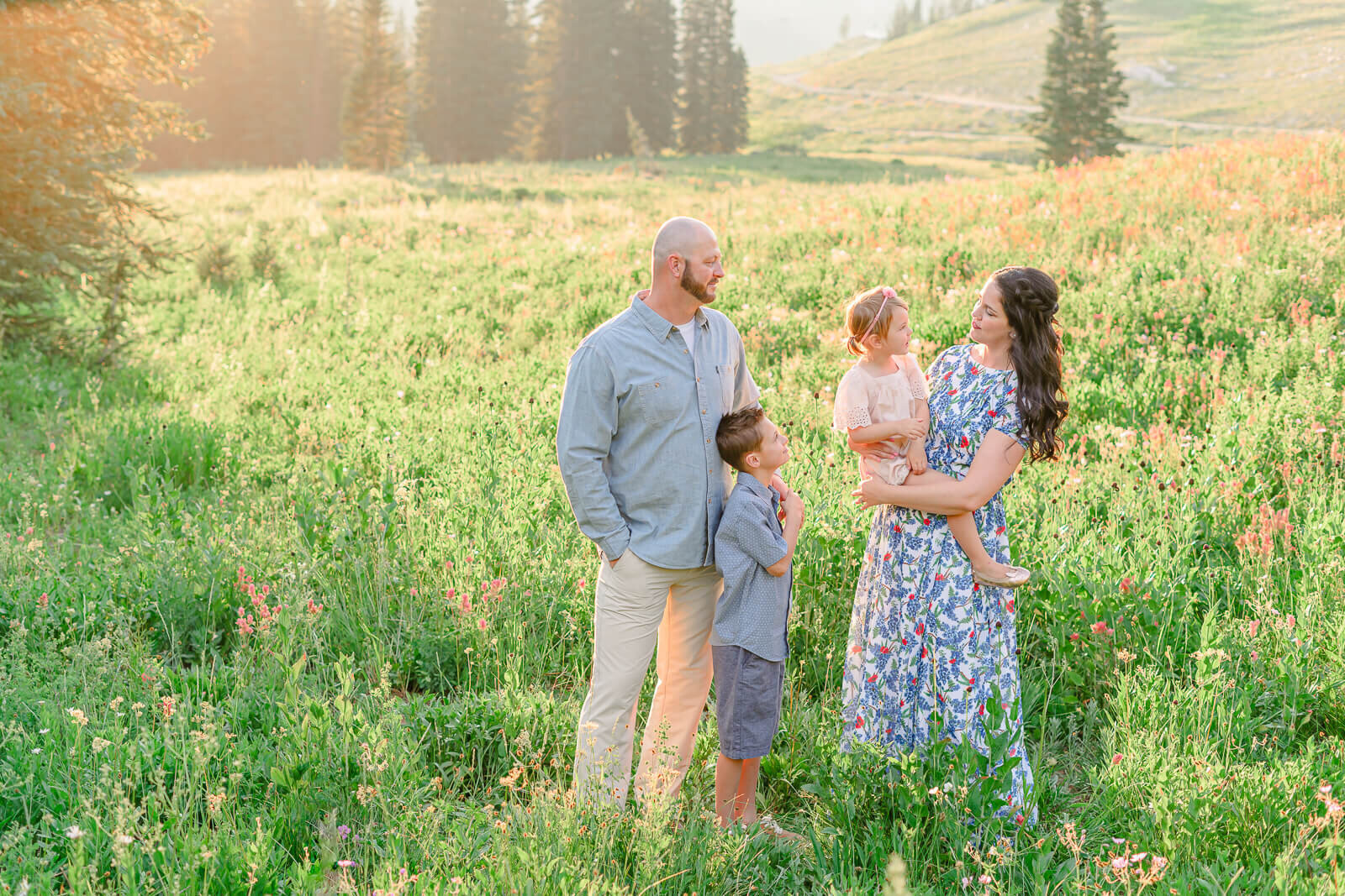 A father, mother, son, and daughter smile and look at each other while standing in a large field of wild flowers at Albion Basin. Captured by Salt Lake Photographer Melissa Woodruff Photography