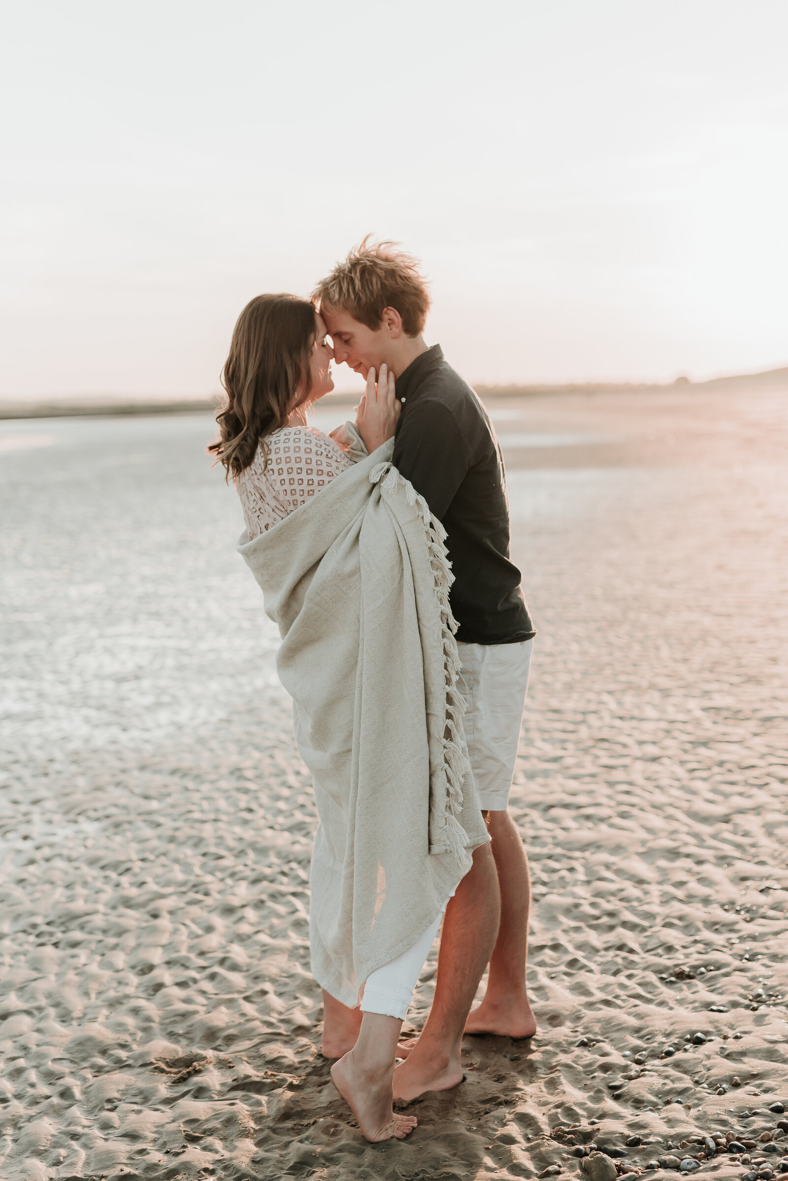 Man embraces his fiance in a cosy cream blanket on a sandy beach as the sun sets