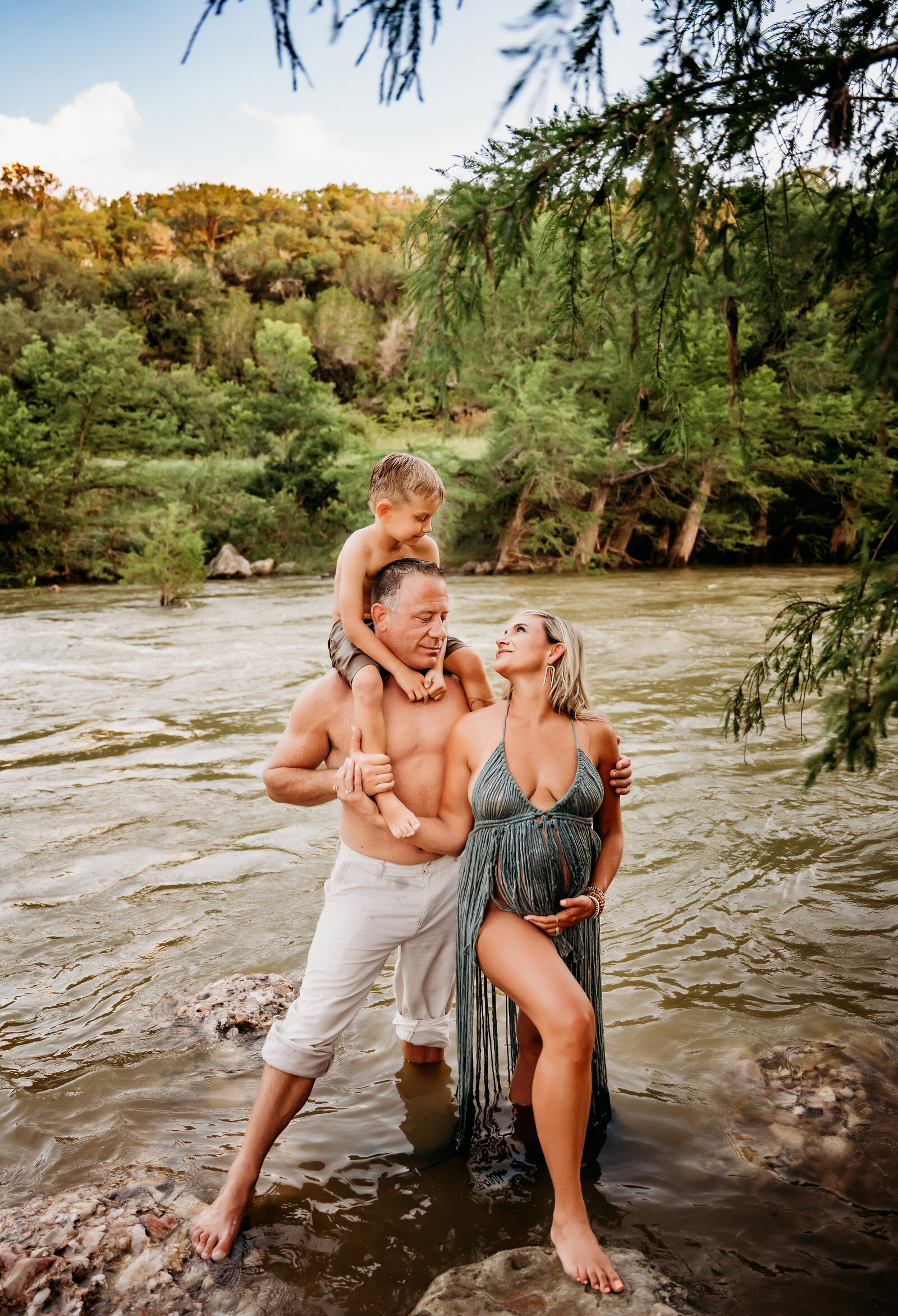 Maternity Photographer, a mother-to-be looks up at her son riding on dad's shoulders at the river