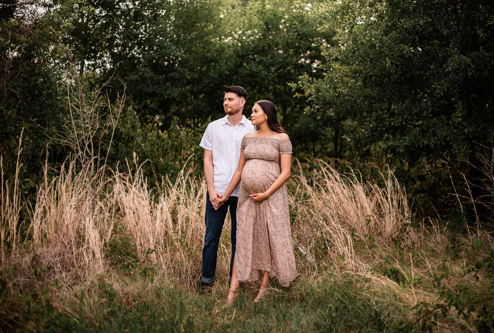 A pregnant couple stand in a field with golden grass and hold hands while looking to their right.