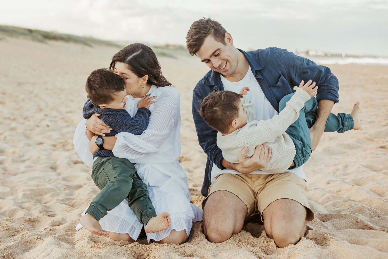 Parents kneeling down and hugging and kissing their little boys at a Sydney beach.