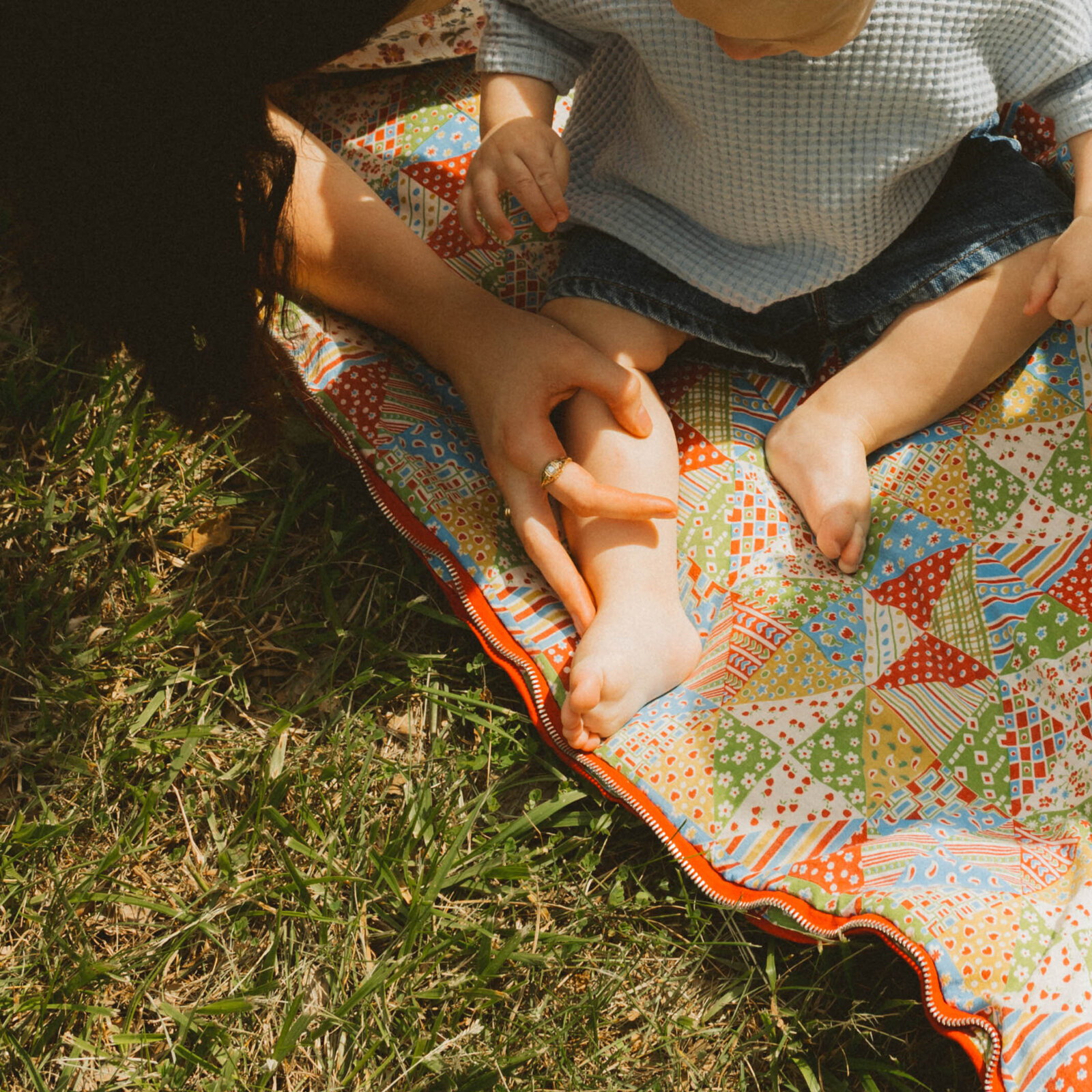 Baby lying on a colorful blanket with legs visible and mother’s arms resting beside him, capturing intimate, tender, and authentic family moment
