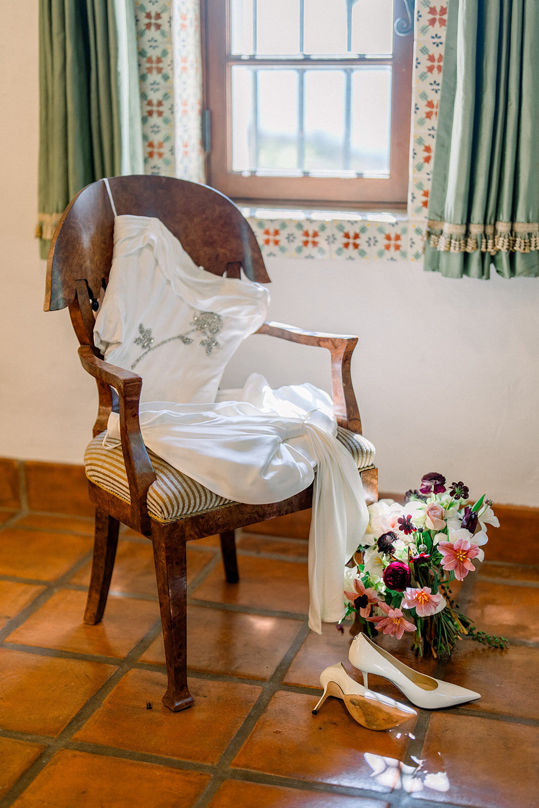 Editorial bridal detail flatlay with bouquet, shoes, and gown arranged on antique chair in bridal suite.