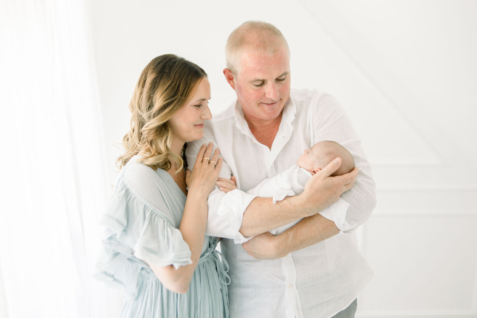 Familienfotografie Vater hält Baby auf dem Arm und Mama hakt sich bei ihm ein und lächelt das Baby an.