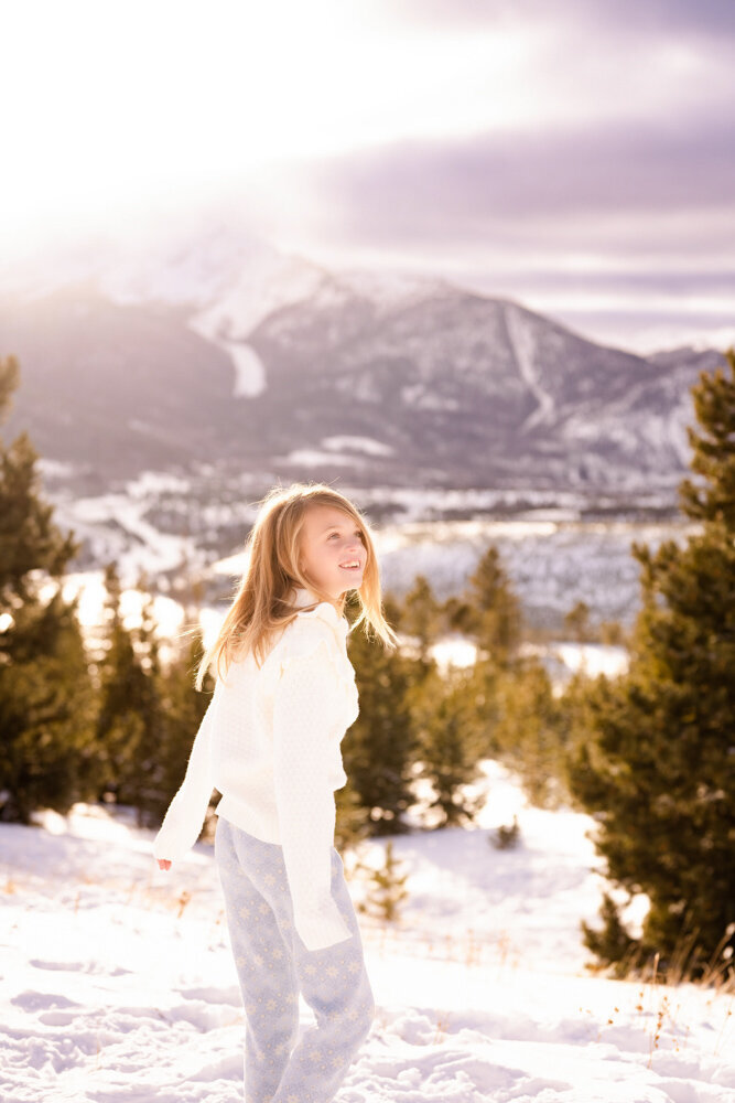 A little girl walks through a forest filled with snow and a mountain backdrop
