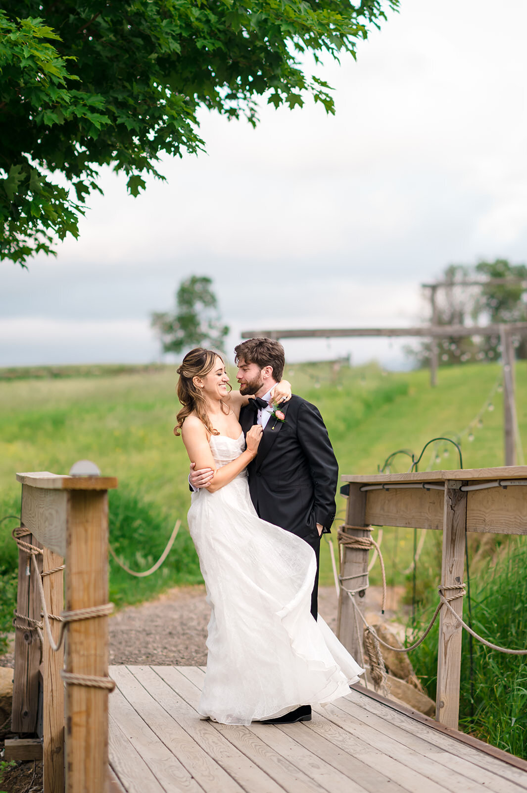 A bride and groom share a laugh on a wooden bridge in a pastoral setting, with the bride playfully grabbing the groom's lapel, surrounded by lush greenery and open skies