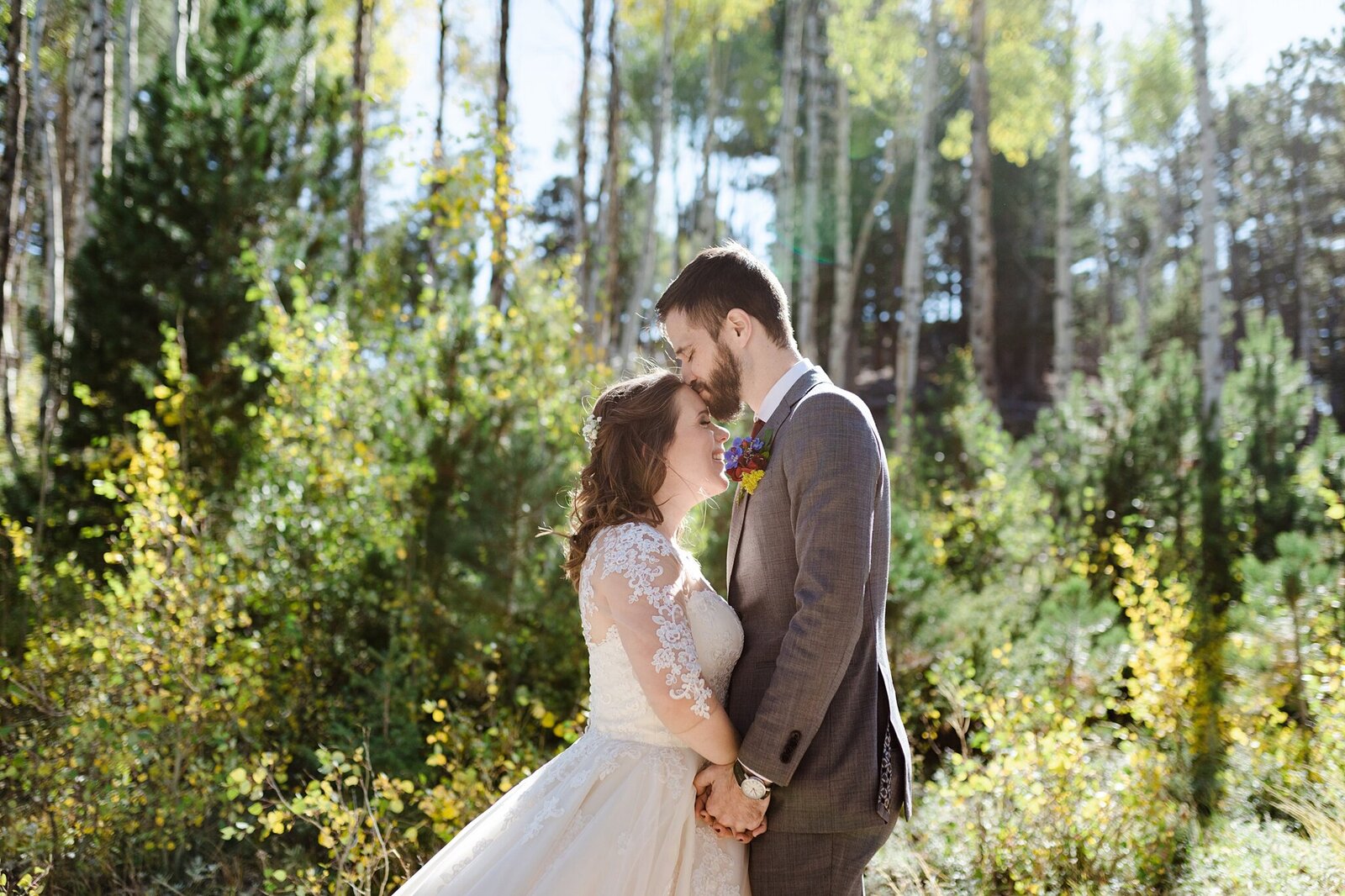 A couple on their wedding day stands in front of aspen trees in Colorado. The bride is smiling, the groom is giving her a kiss on the forehead, and they are holding hands.