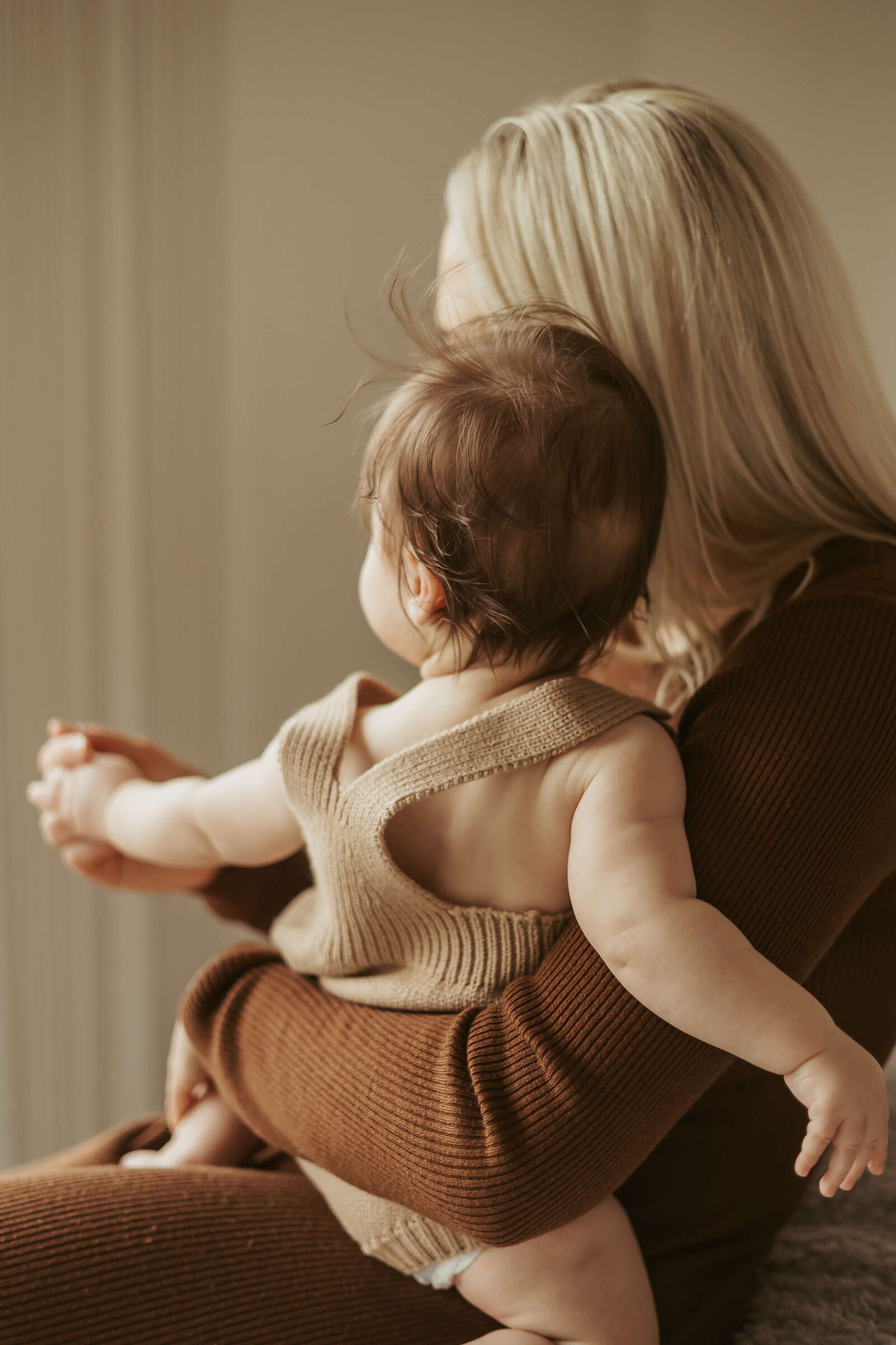 A beautiful mum with blonde hair and wearing a long-sleeve brown dress is holding her baby while they look out the window.
