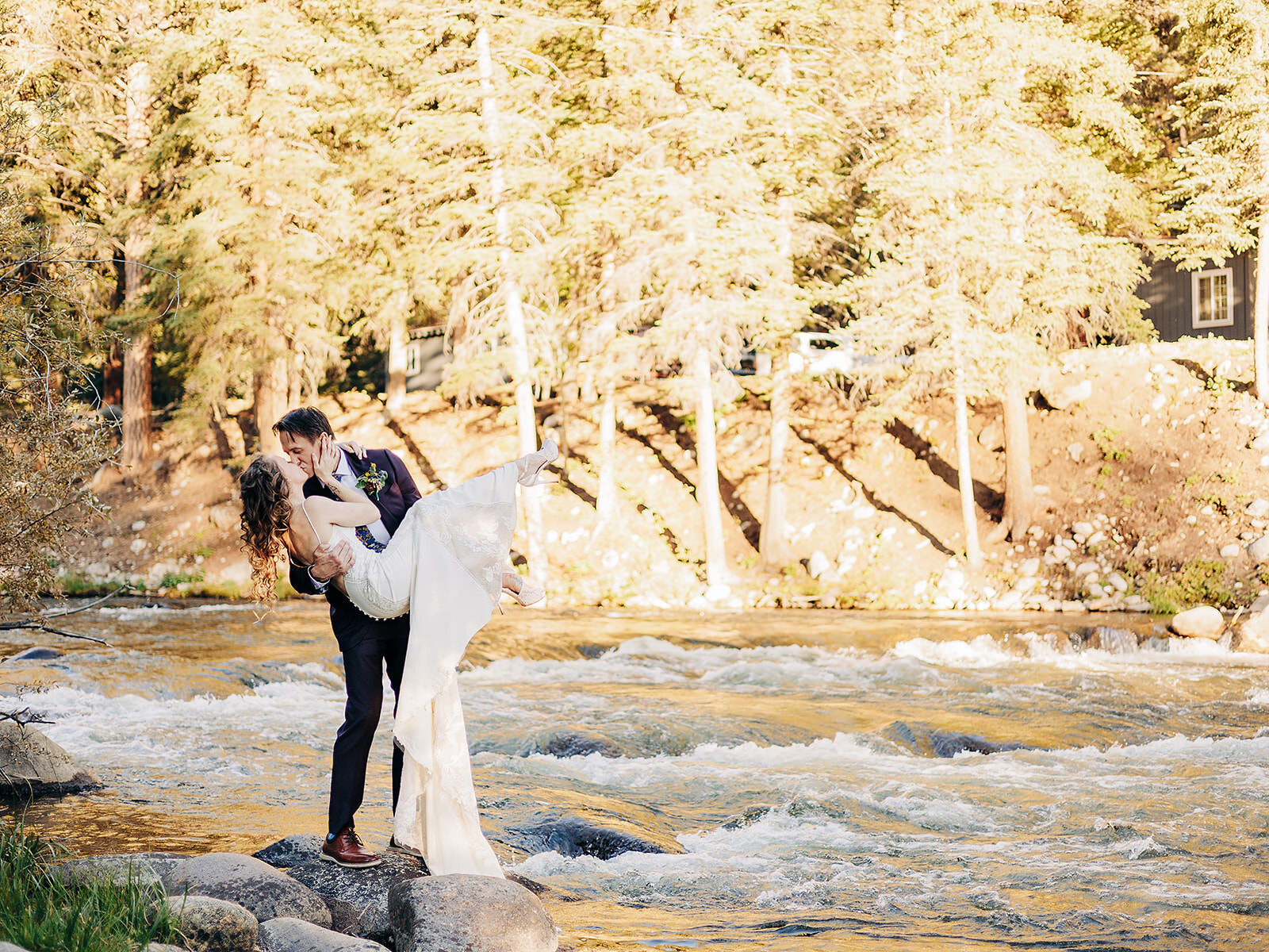 A groom lifts and kisses the bride beside a flowing river, surrounded by rocks and sunlit trees, as a Colorado Wedding Photographer captures this romantic and joyful outdoor wedding scene.