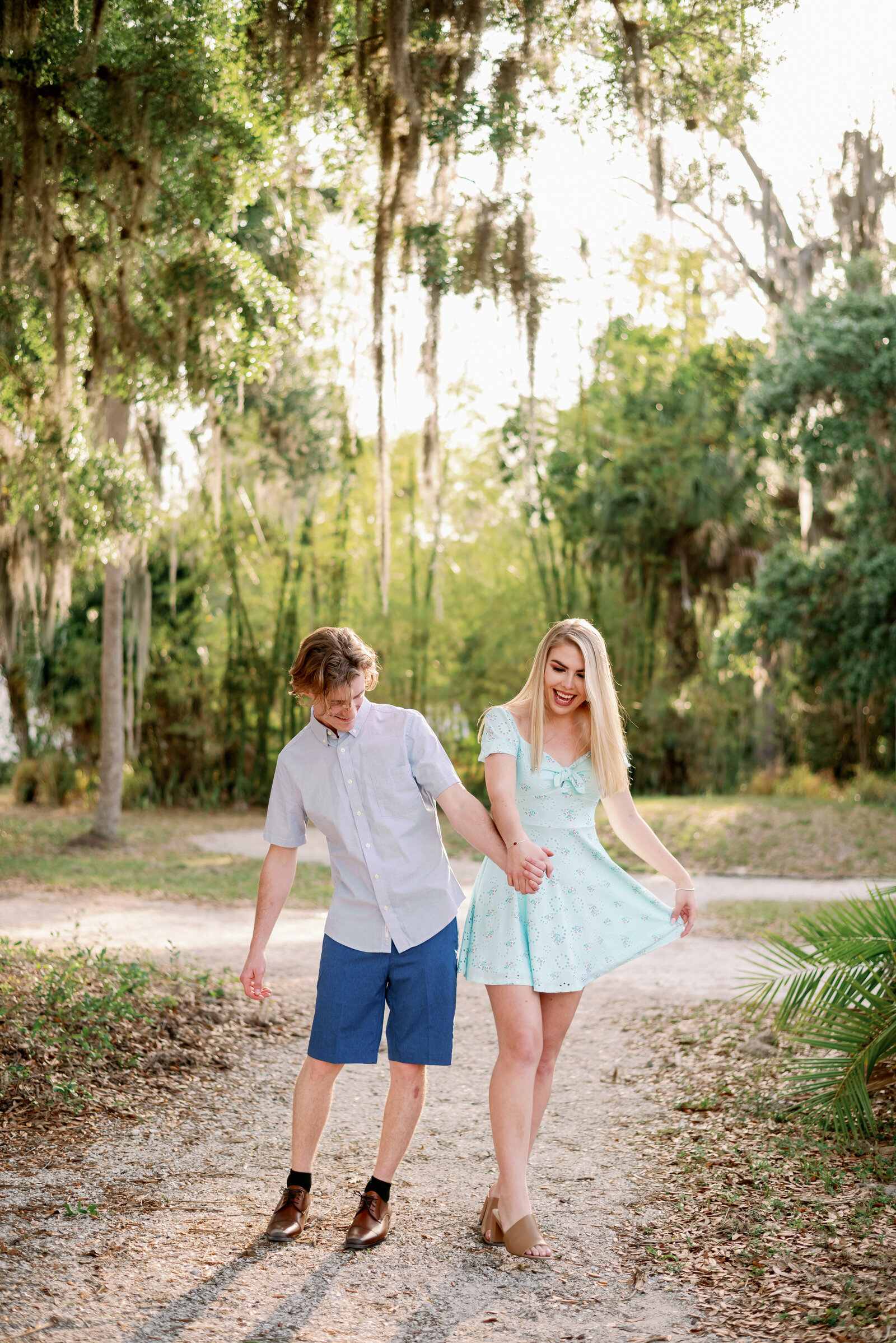 engaged couple wallking down a path toward the camera trying to bump their hips together as they walk. Both are laughing and holding hands
