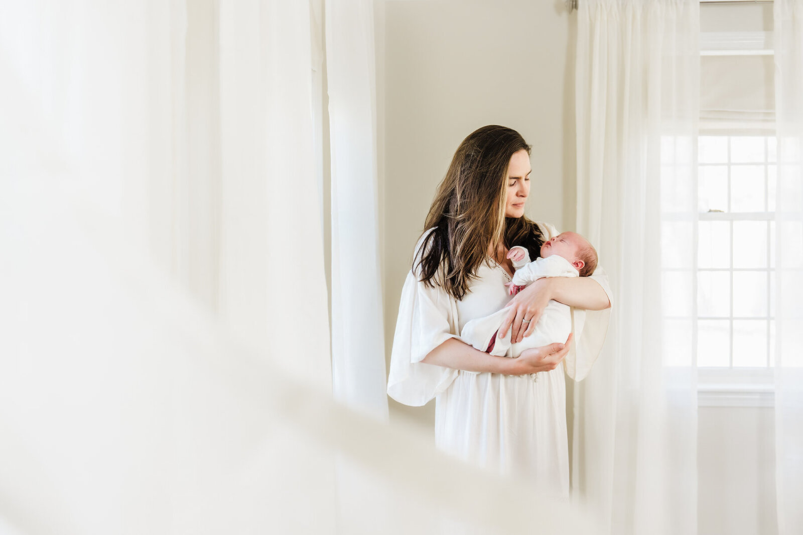 mother in white dress stands in white room gazing at infant boy