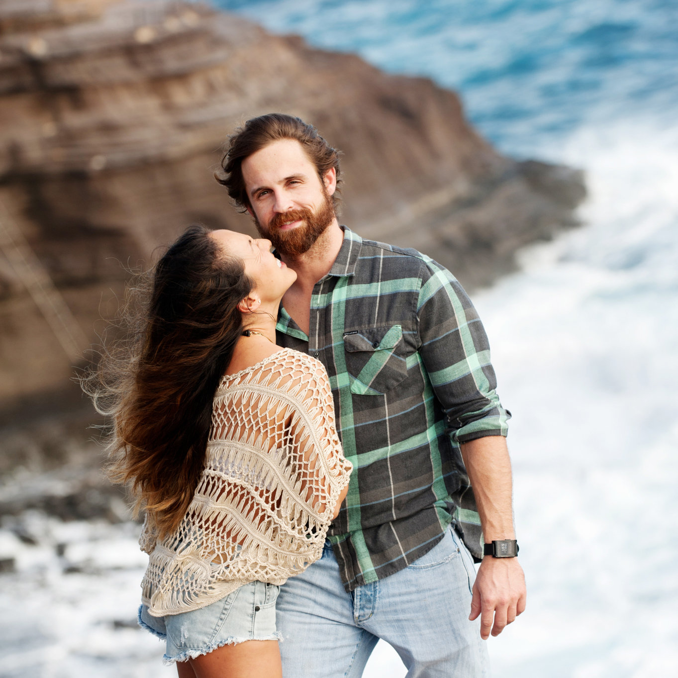 a handsome man looks at the camera as a woman reaches up to give him a kiss with spitting caves hawaii in the background