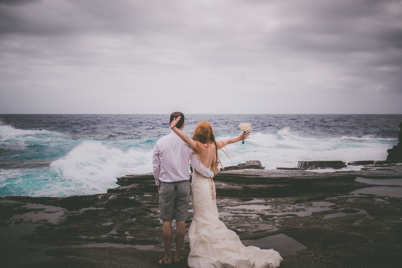 Bride raises arms when wave crashes in Hawaii.