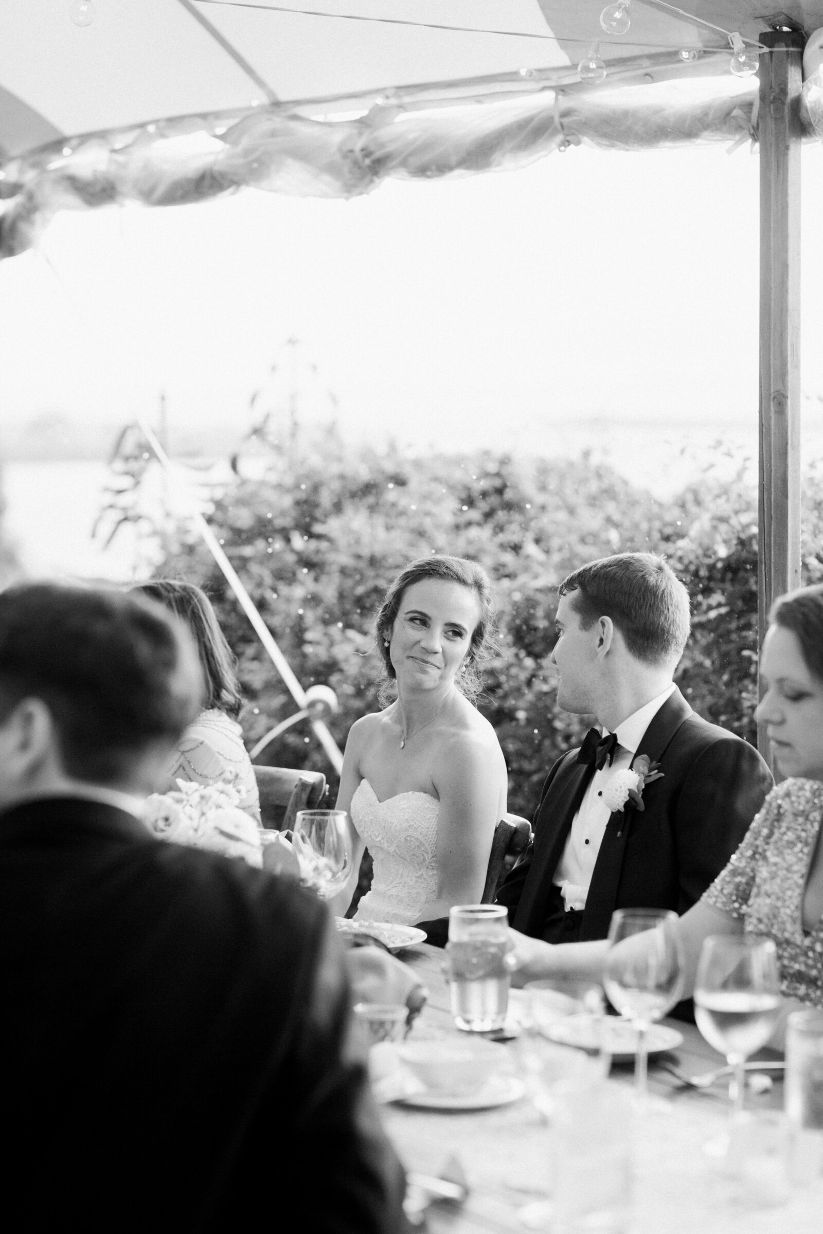 Black and white photo of a couple sitting at a table at their wedding reception with their guests.
