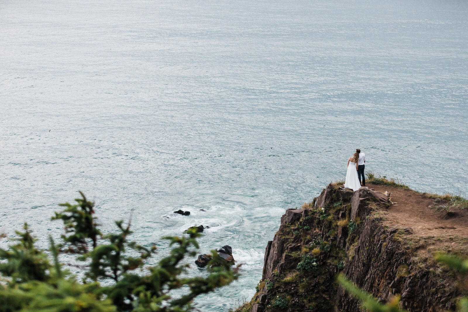 A blonde bride wearing a strapless gown holds her groom's hand and rests her head on his shoulder as they gaze out at the Pacific Ocean while standing on the cliff edge after saying their vows at their Oregon Coast adventure elopement near Manzanita. | Erica Swantek Photography