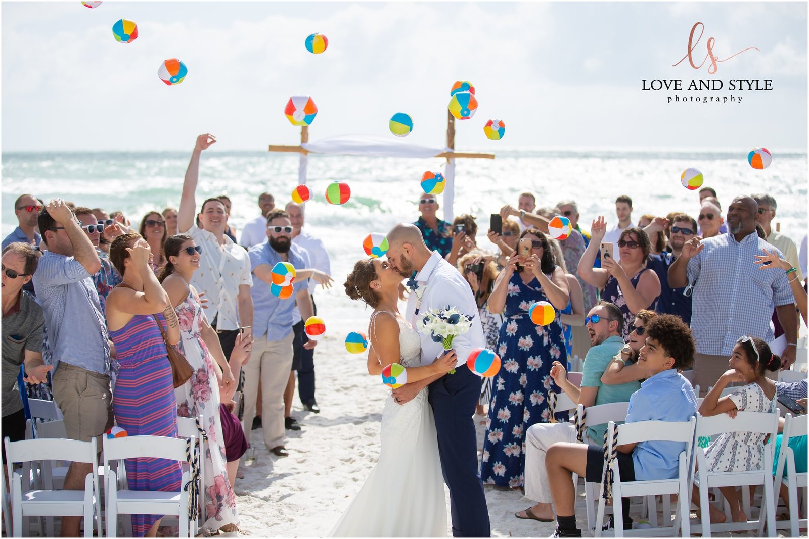Bride and Groom kissing at the end of the aisle while guests throw beach balls in the air at The Beach House Restaurant on Anna Maria Island