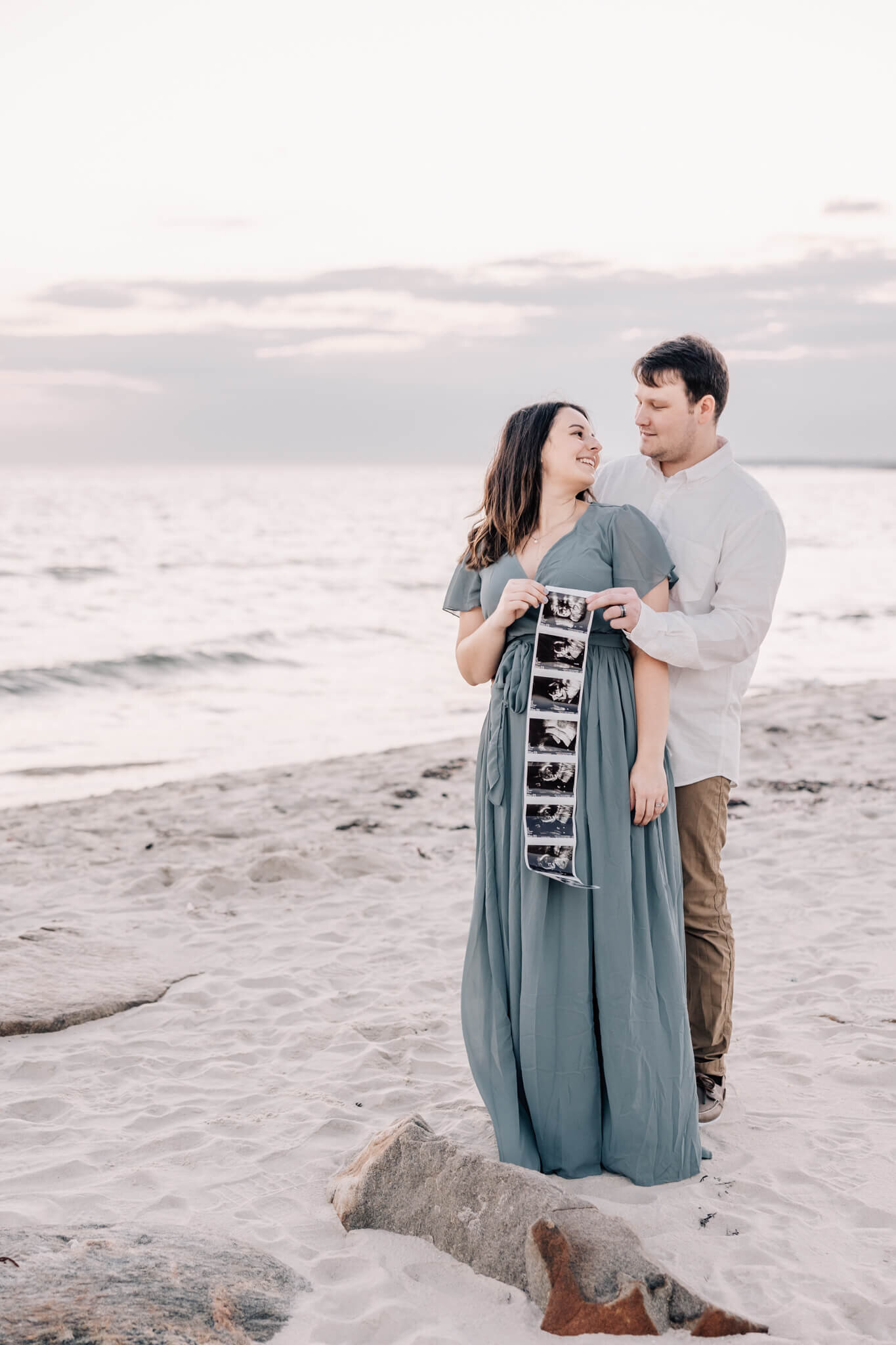 husband and wife look at each other at beach holding ultrasound photos