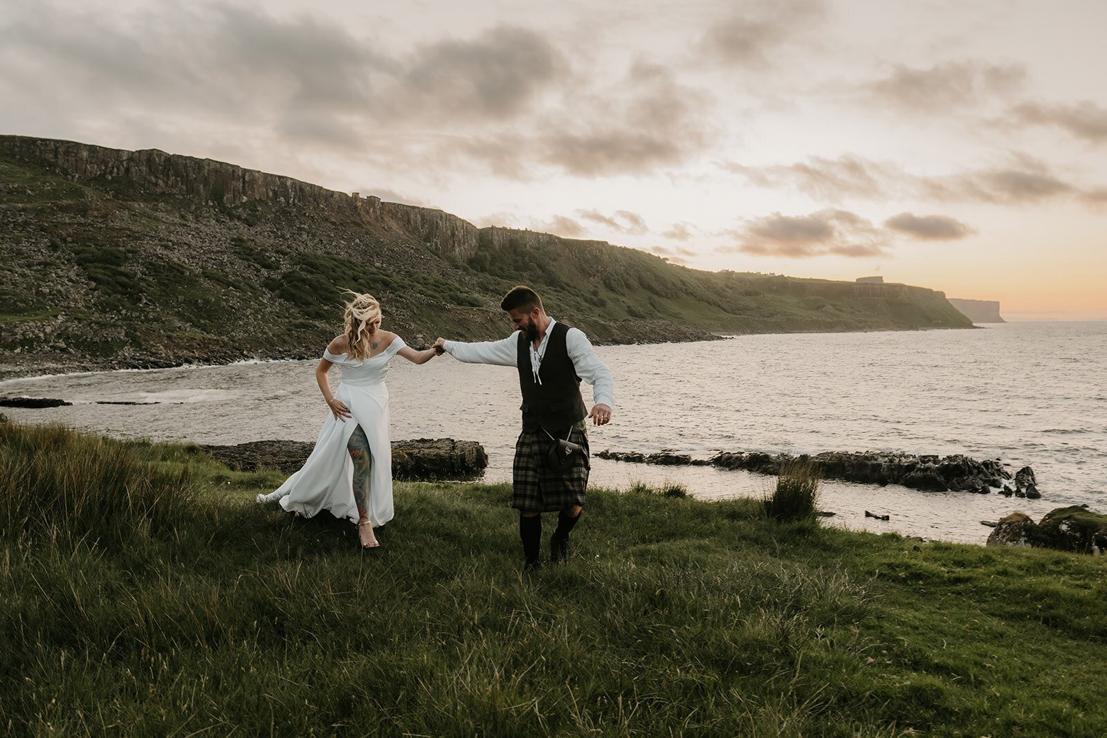 A couple, dressed in wedding attire, walk hand-in-hand by the sea with cliffs in the background at sunset—a serene moment perfect for a California photography retreat for storytellers and soul seekers.