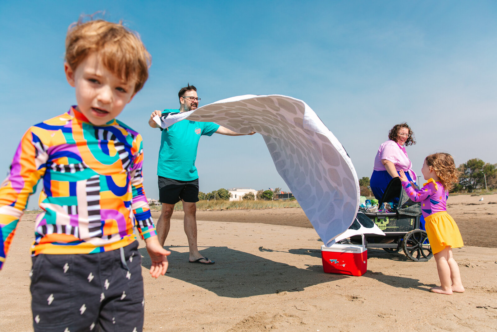photo of a family at the beach with dad spreading out a blanket and the rest of the family unpacking