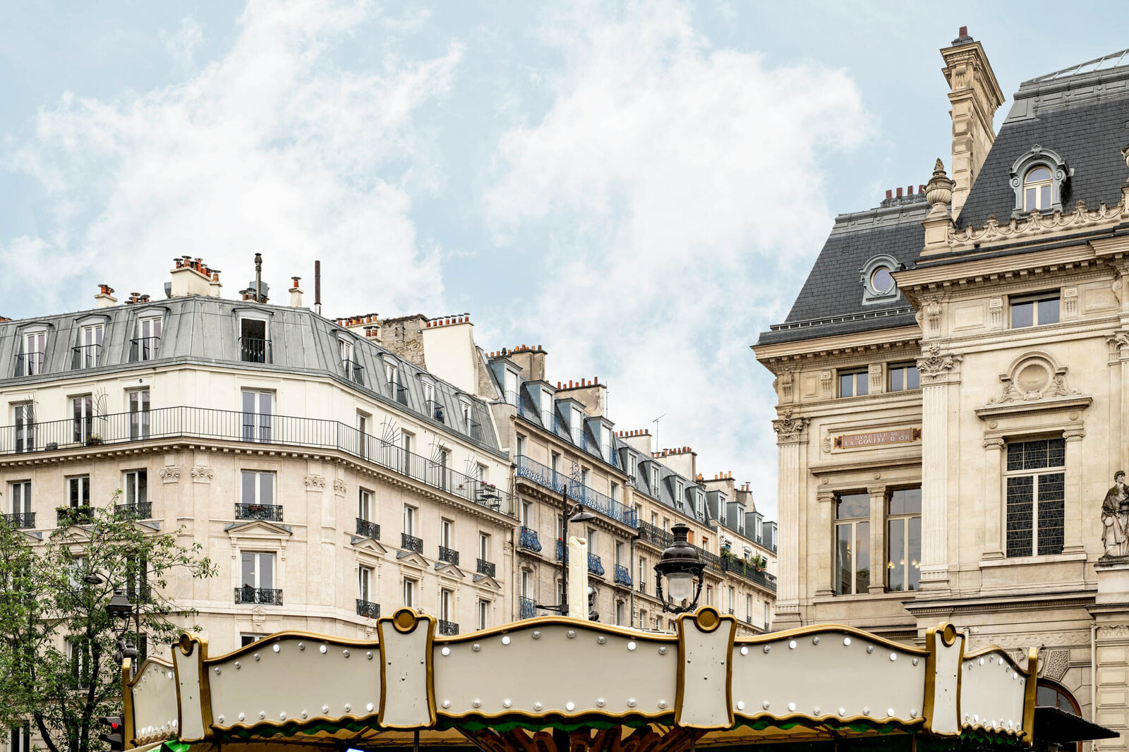 A Parisian streetscape features classic Haussmann-style buildings with mansard roofs and ornate facades. In the foreground, a vintage carousel top is partially visible under a blue sky with scattered clouds.
