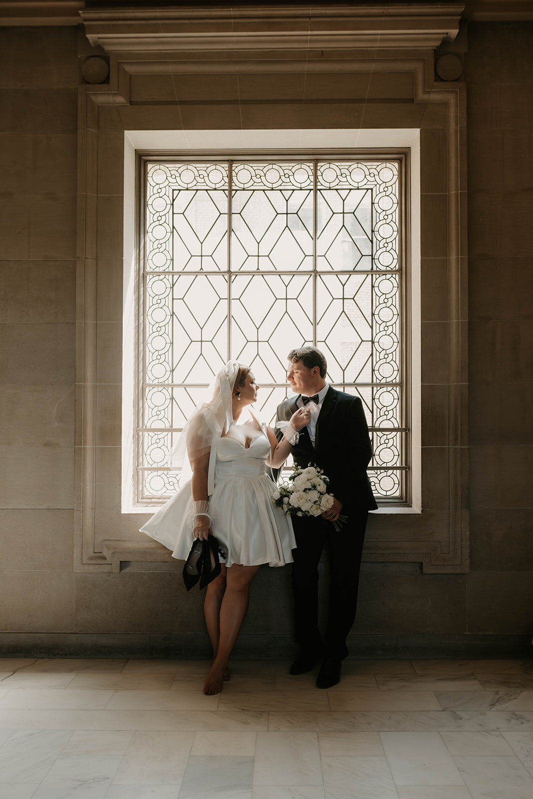 Bride and groom during sunset elopement stand in front of a sunlit ornate window, smiling at each other and holding flowers.