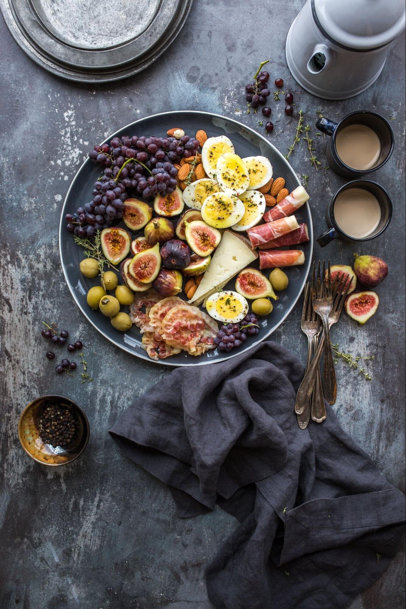 Image: boiled egg halves, figs, green olives, and purple table grapes with procusto and a cheese wedge on a black plate