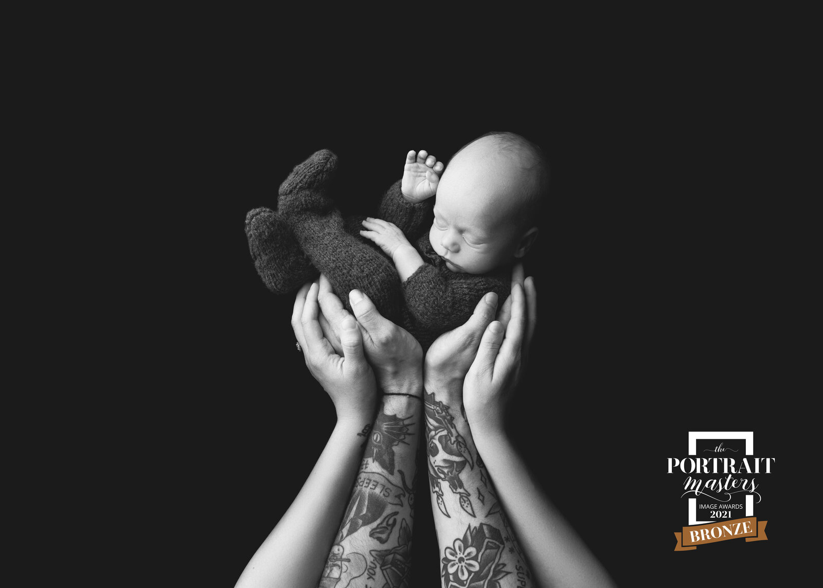 black and white image of parents holding up their newborn; dad has tattooed arms
