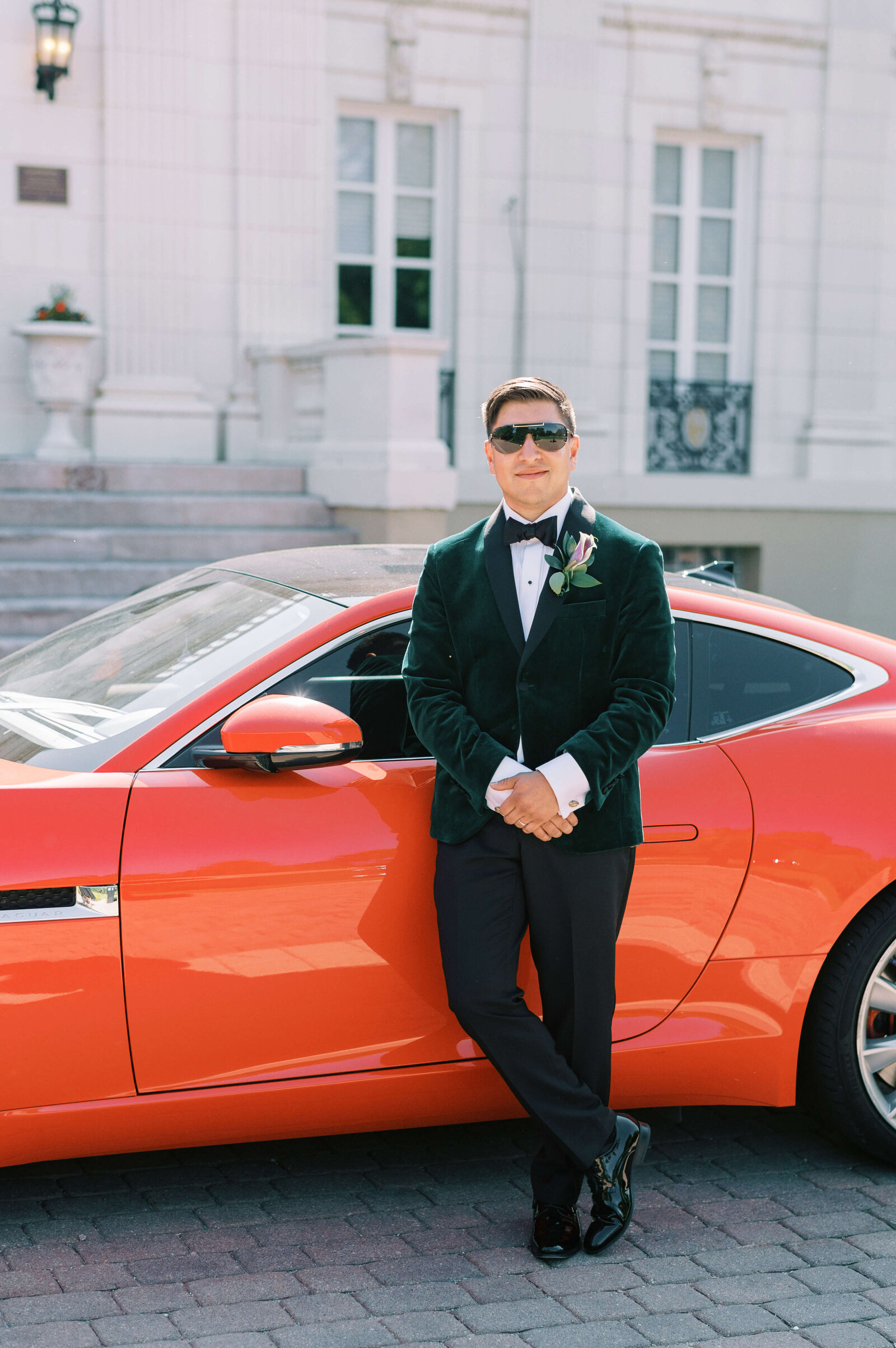 a handsome groom in an emerald suite stands in front of a  red sports car
