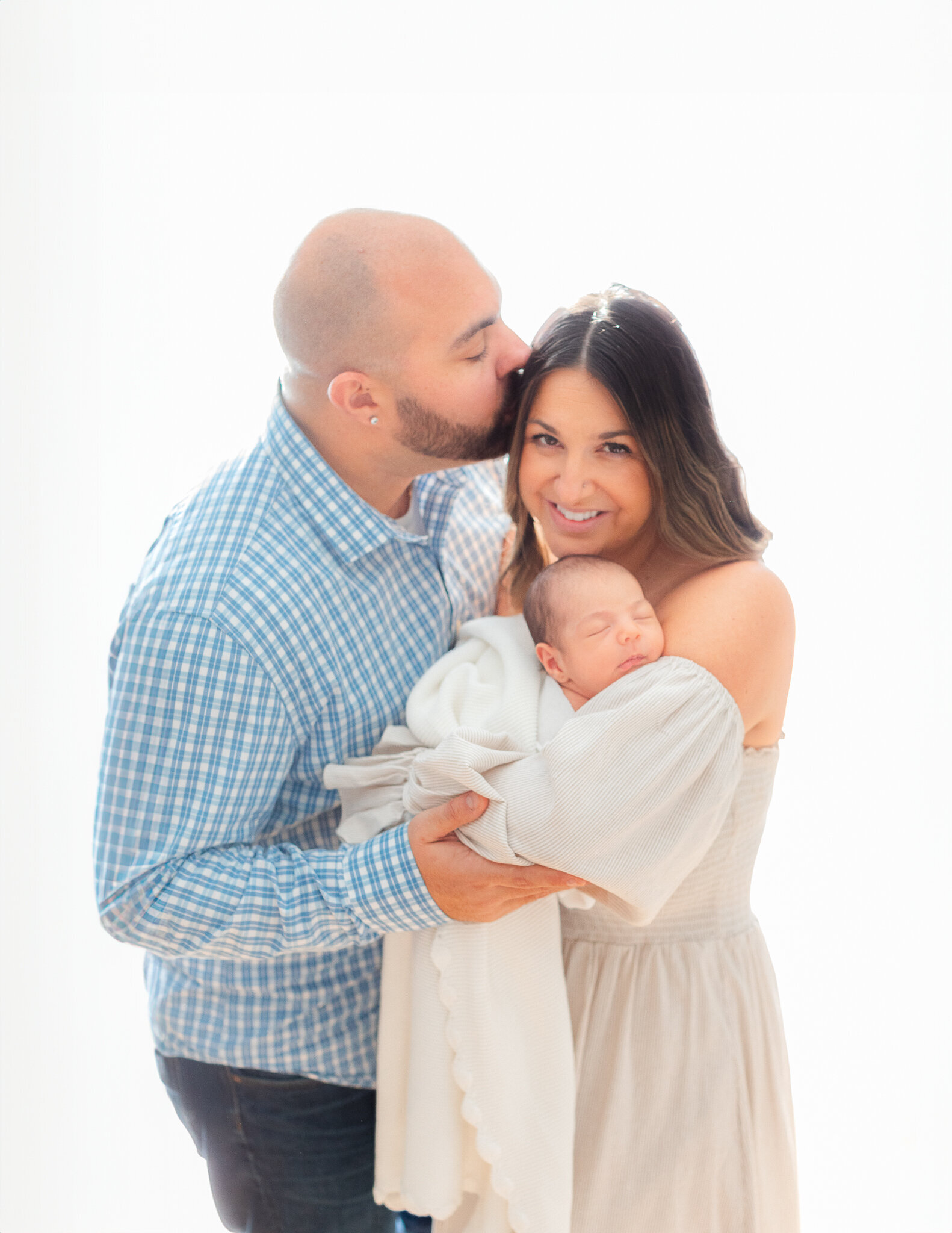 mom wearing dress smiles at camera holding newborn baby while dad kisses her head during massachusetts newborn photo session