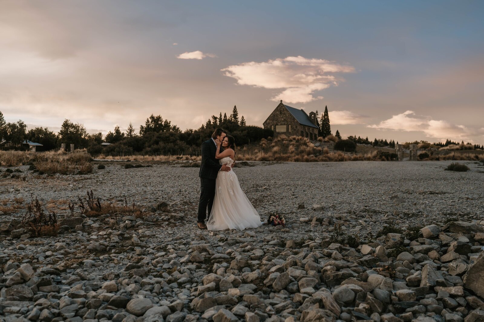 Bride and groom embrace on a rocky landscape with a rustic church in the background at sunset, capturing the magic of an Italy photography retreat in the countryside for inspired storytelling.