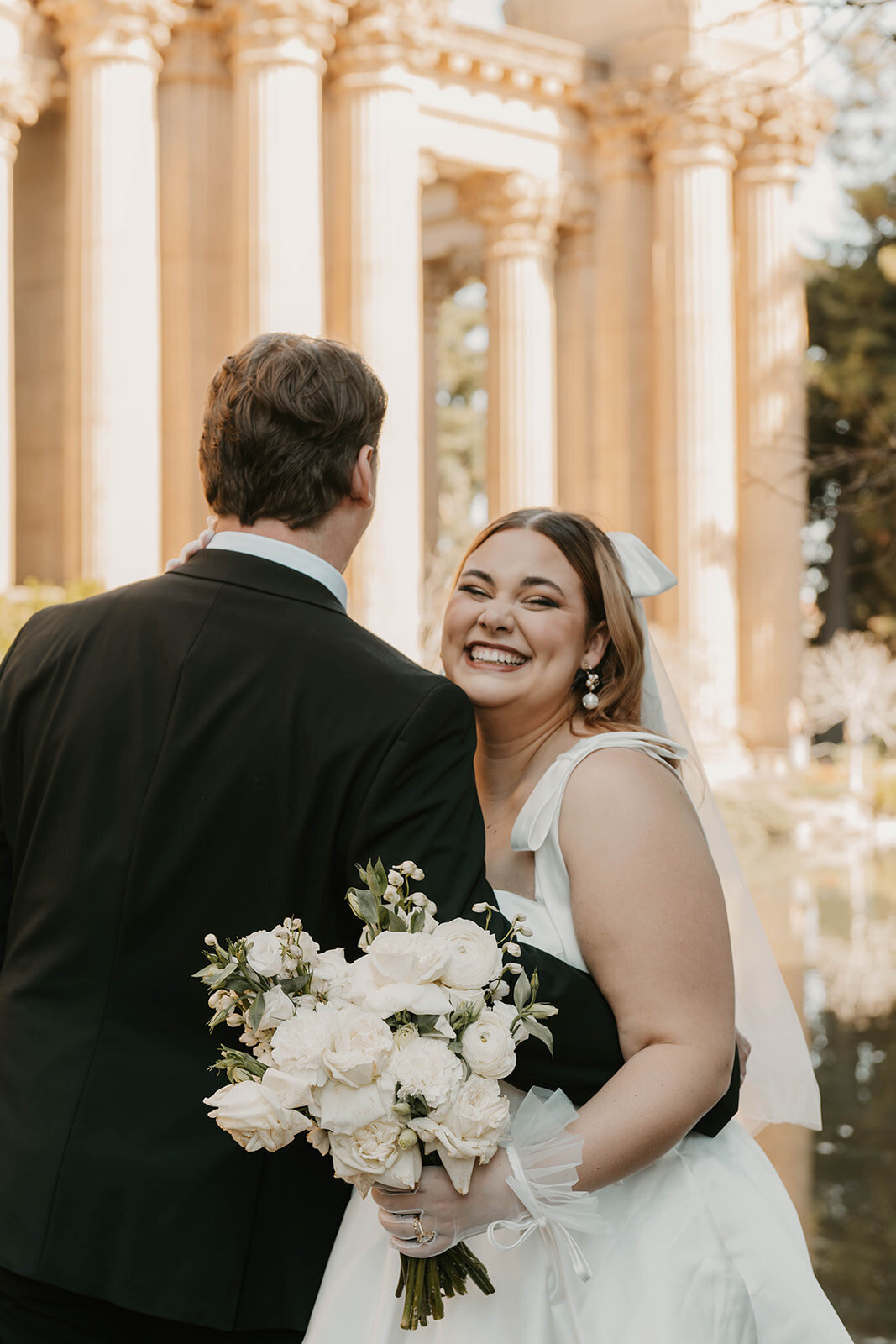 The bride smiles and holds a bouquet, standing next to the groom outdoors in front of grand columns during their Colorado wildflower elopement.