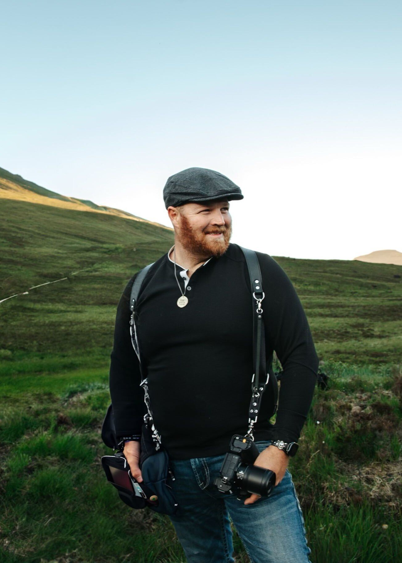 Man with a beard wearing a flat cap and camera gear stands outdoors in a grassy, hilly landscape behind the scenes at creative photography retreat.