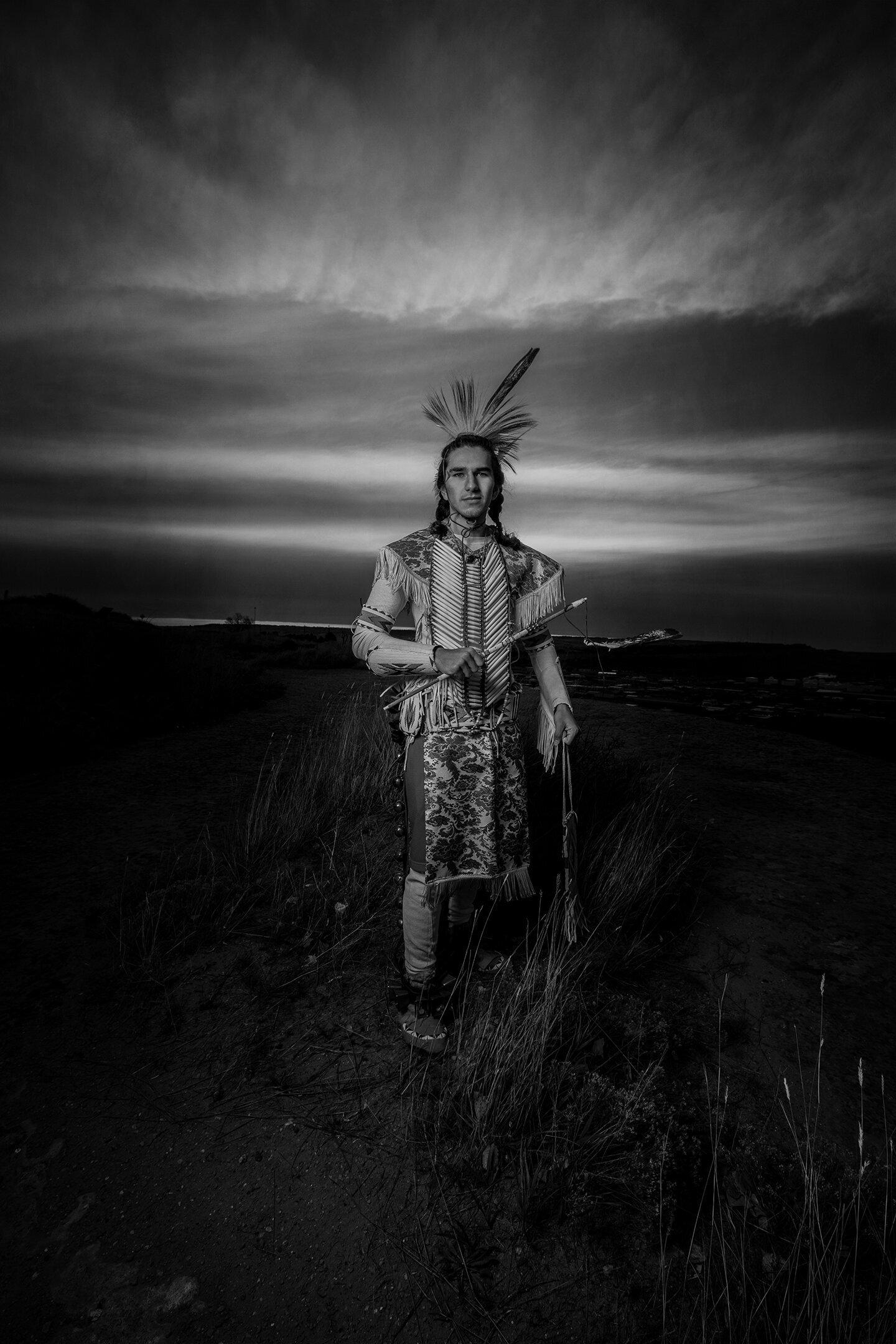 Black and white picture of Native American in tradition regalia. Senior pictures on the rims in Billings MT. Feathers, headdress, beading. Prairie grass.