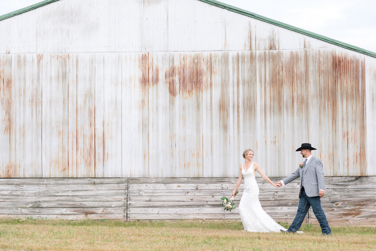 bride and groom walking in front of white aged metal barn on family owned steer breeding land