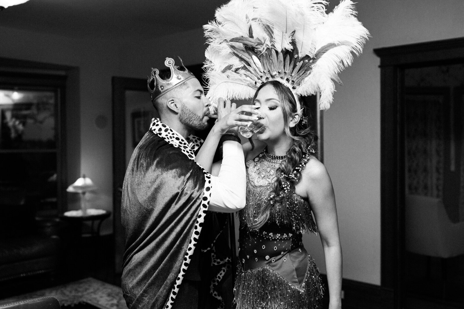 A man in a crown and royal cape drinks from a glass held by a woman in a feathered headdress and decorative dress. Captured by a Colorado Wedding Photographer, they stand close together in festive costume attire in a cozy living room.



