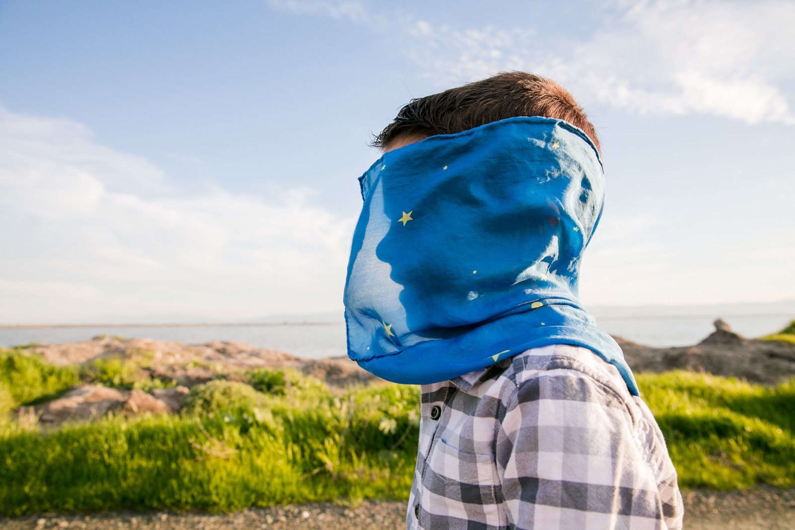 photo of a young boy in plaid shirt standing in front of a blue sky and green hill with a blue play silk being blown in the wind and covering his face with his profile backlit and showing through the fabric