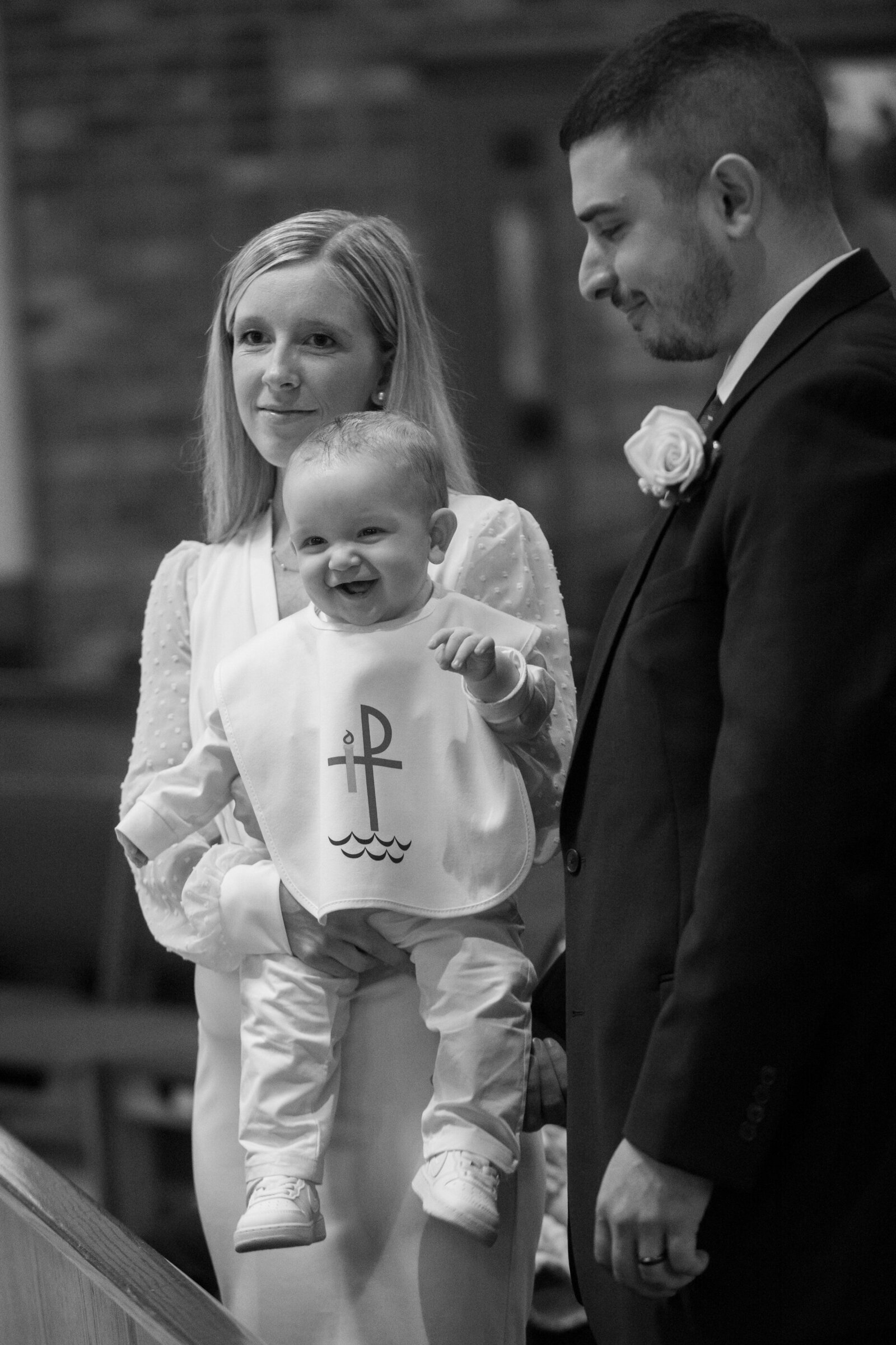 Black and white photo of a white mom holding an 8-month-old baby wearing baptism bib stands with father in a dark suit with flower at Saint Augustine New City Rockland County baptism photographer Milestone Images