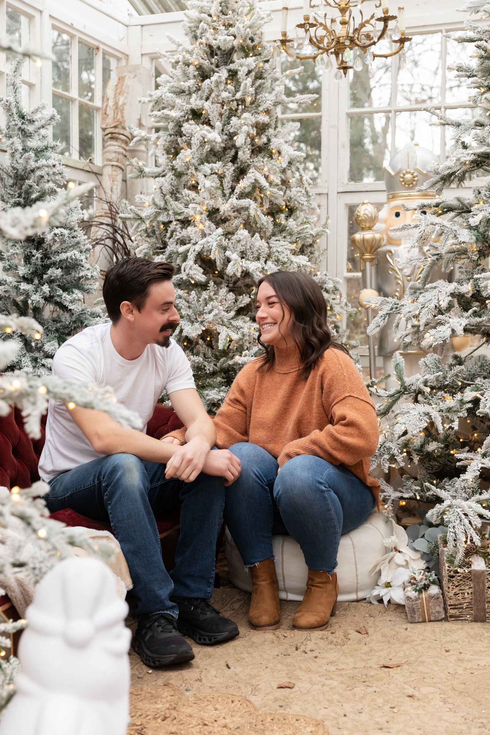 couple sits on sofa and pouf, looking at each other laughing in greenhouse decorated for christmas at rustic wild arrow in brooklyn michigan