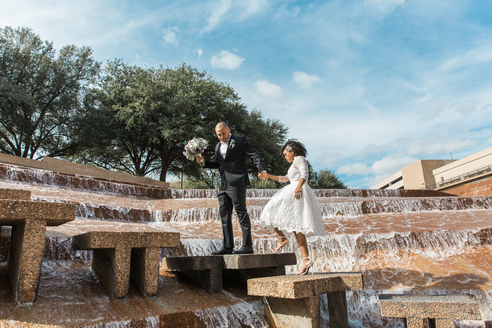 A groom leading a bride across the steps of an intricate water fountain at the Fort Worth Water Gardens in Fort Worth Texas. The bride is on the right and is wearing a short white lace dress with white heels. The groom is on the left and is wearing a black tuxedo with a boutonniere and is holding a bouquet. The steps they're on are surrounded by many levels of waterfalls.