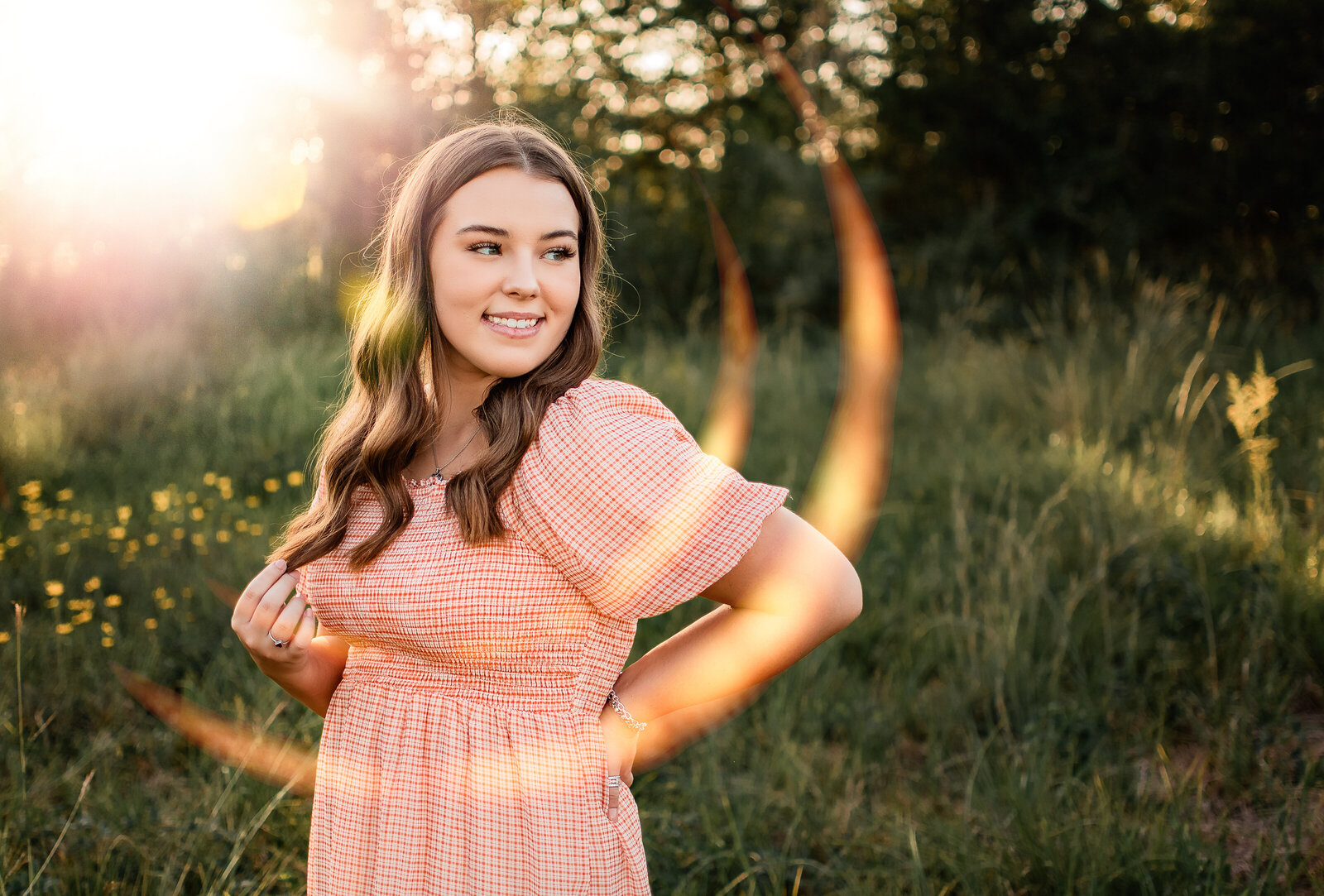 A high school senior stands in a field and smiles over her shoulder as the sun shines on her.