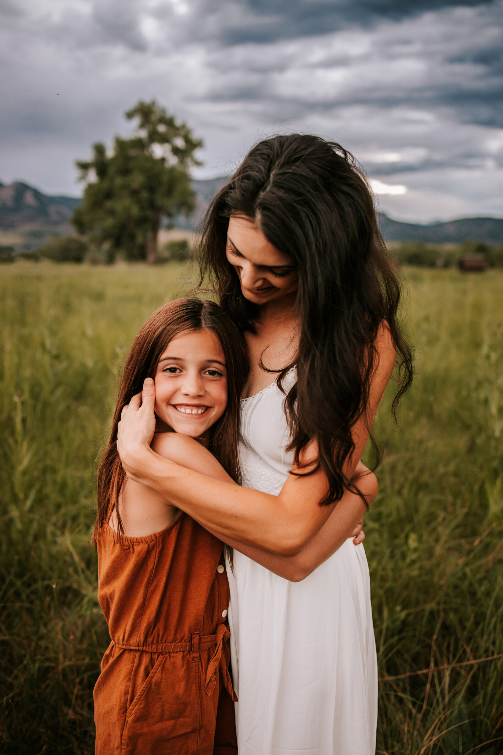 mom hugs young daughter in  the middle of a grassy field while wearing an orange and white dress during golden hour