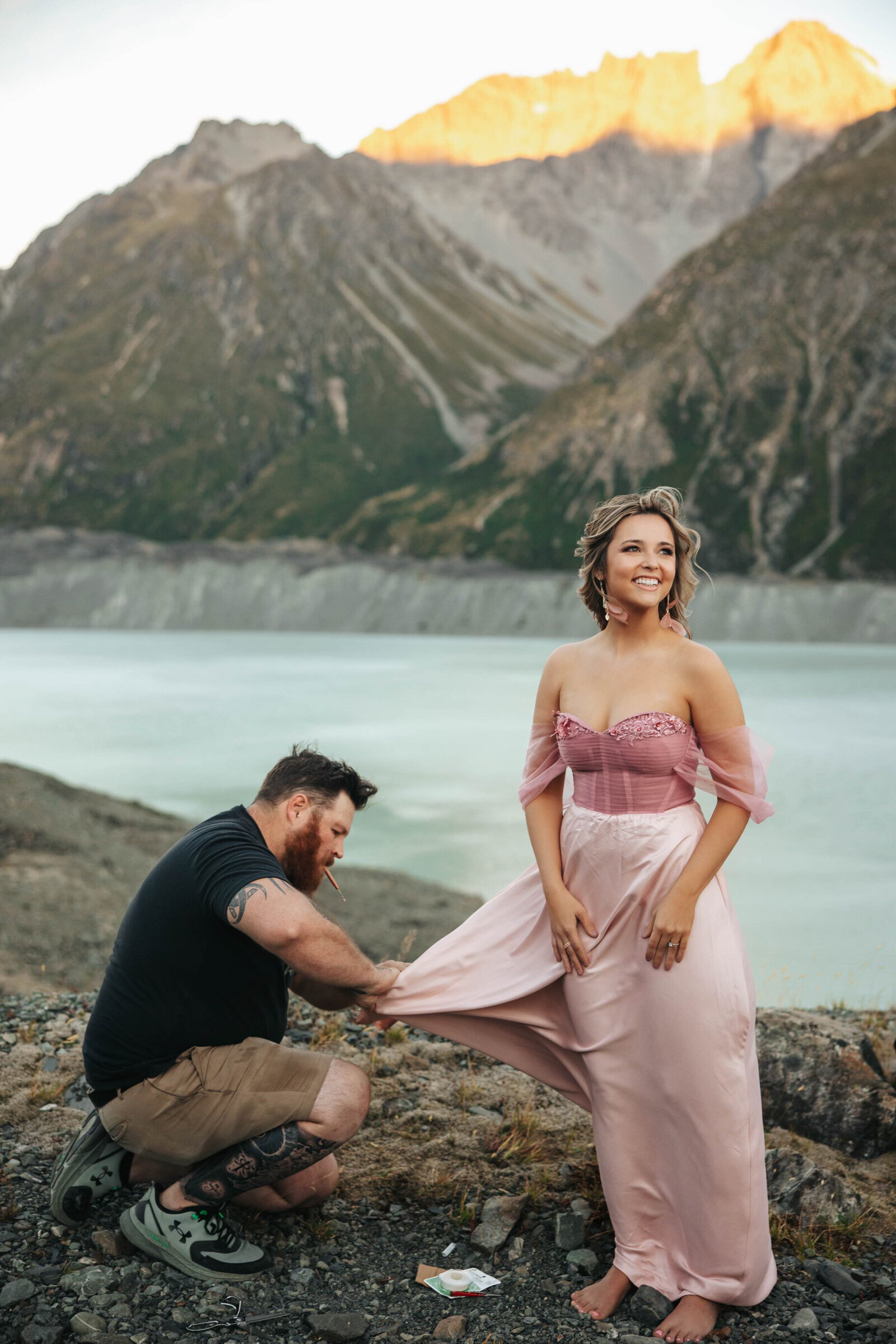 A man adjusts a woman's pink dress outdoors by a lake with mountains in the background, evoking the spirit of an Oregon coast photography retreat for wild-hearted artists.