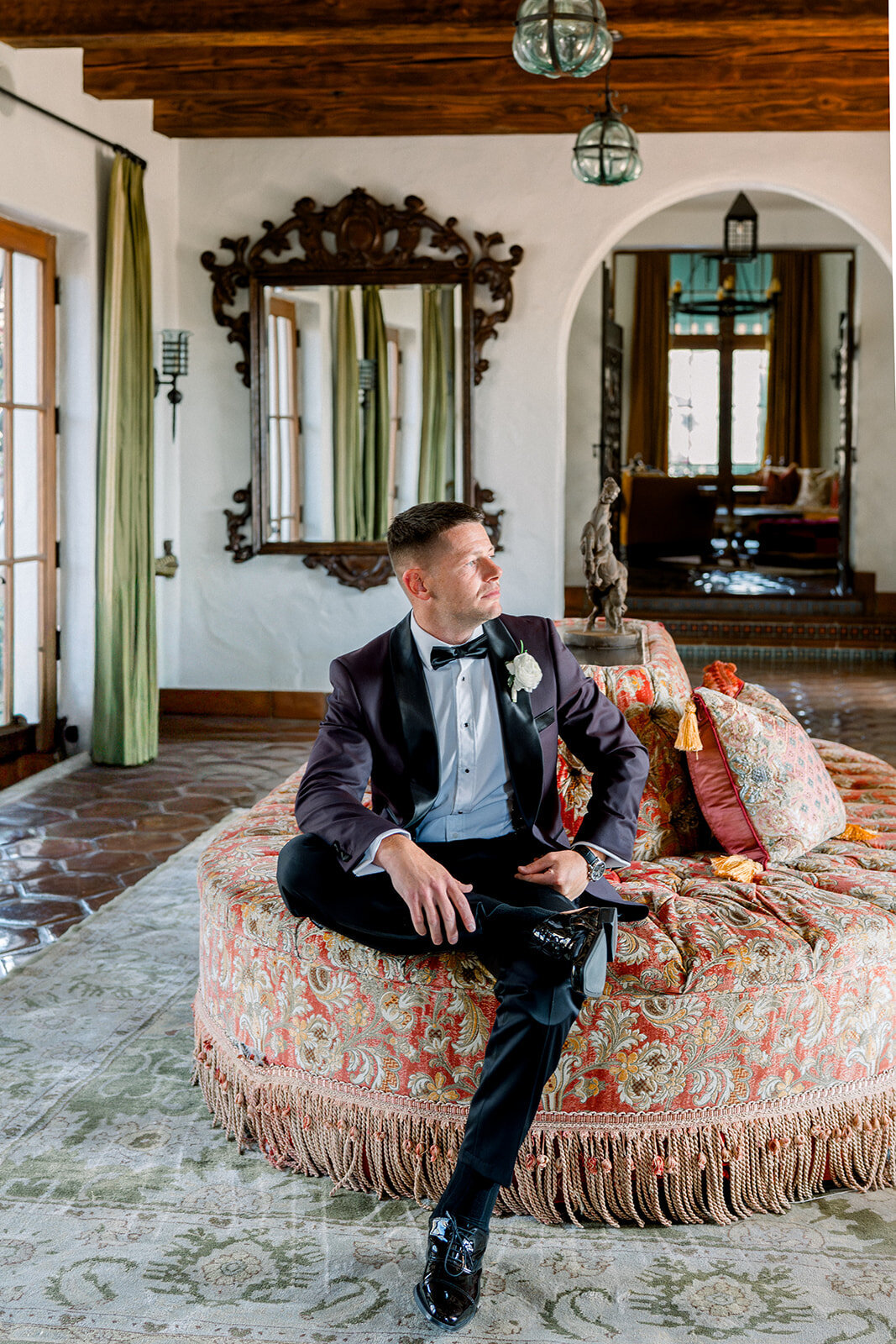 Groom seated confidently in ornate bridal suite with antique furniture, captured in editorial wedding photography style.