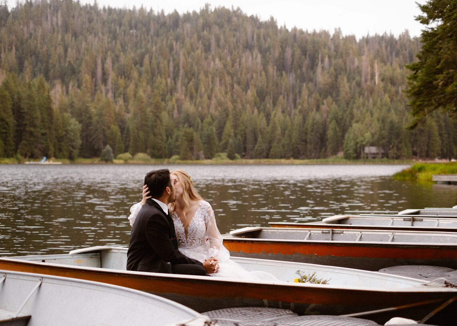 Bride and groom sharing a kiss while sitting in a metal rowboat on a quiet mountain lake, surrounded by pine trees.