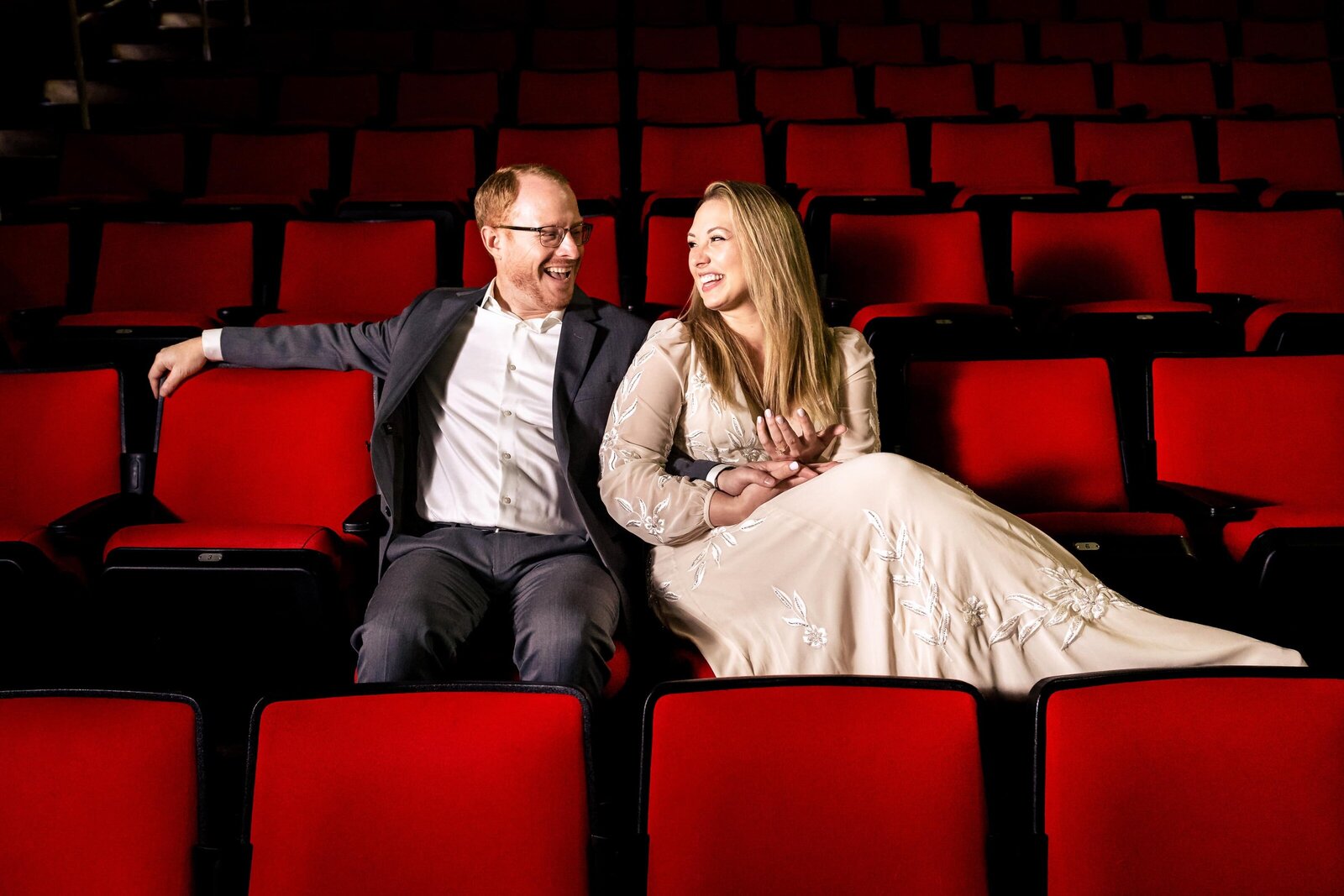 man in a suit and woman in a white dress sit next to one another in a row of red stadium seats