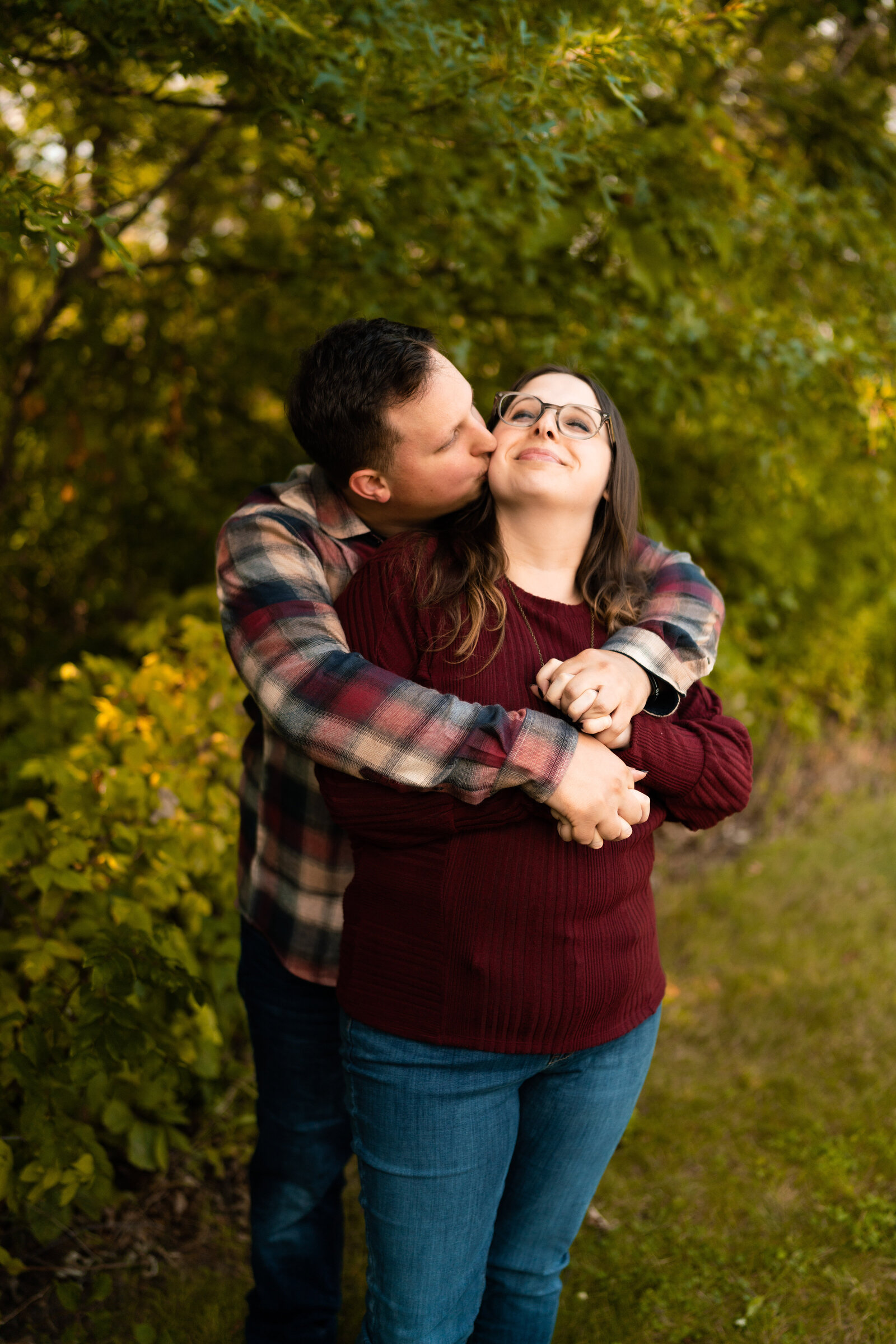 Man in flannel and woman in red sweater snuggle and kiss in park.
